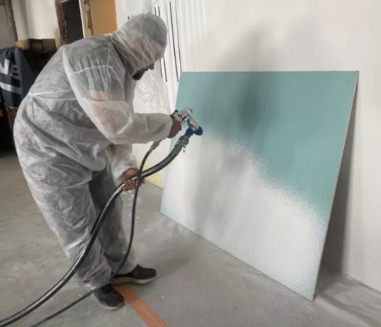 Pracownik w kombinezonie maluje płytę kartonowo-gipsową natryskowo, od dołu na biało, góra płyty w kolorze miętowym, widoczny pistolet natryskowy i węże.