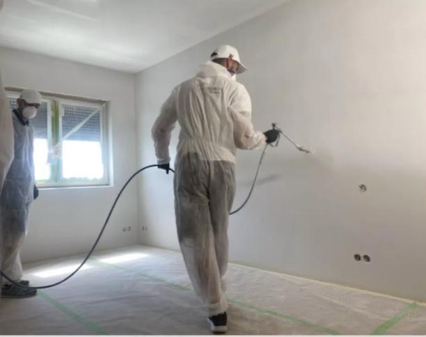 Malowanie ścian natryskowo przez dwóch pracowników w kombinezonach ochronnych. Podłoga zabezpieczona folią malarską.