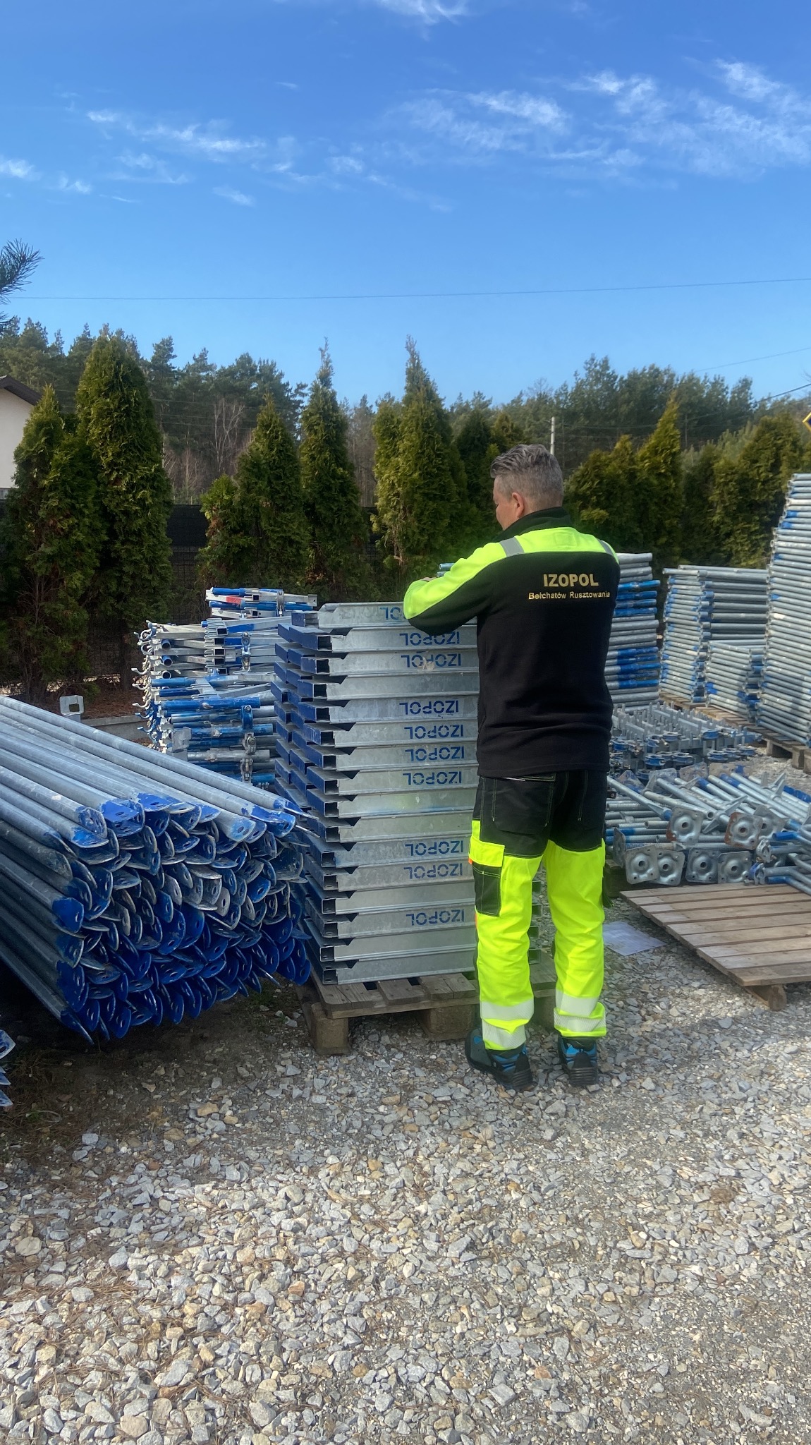 Pracownik w odzieży roboczej firmy Izopol stoi obok stosu elementów rusztowań na żwirowym podłożu, w tle iglaki i błękitne niebo. Widoczne elementy rusztowań.