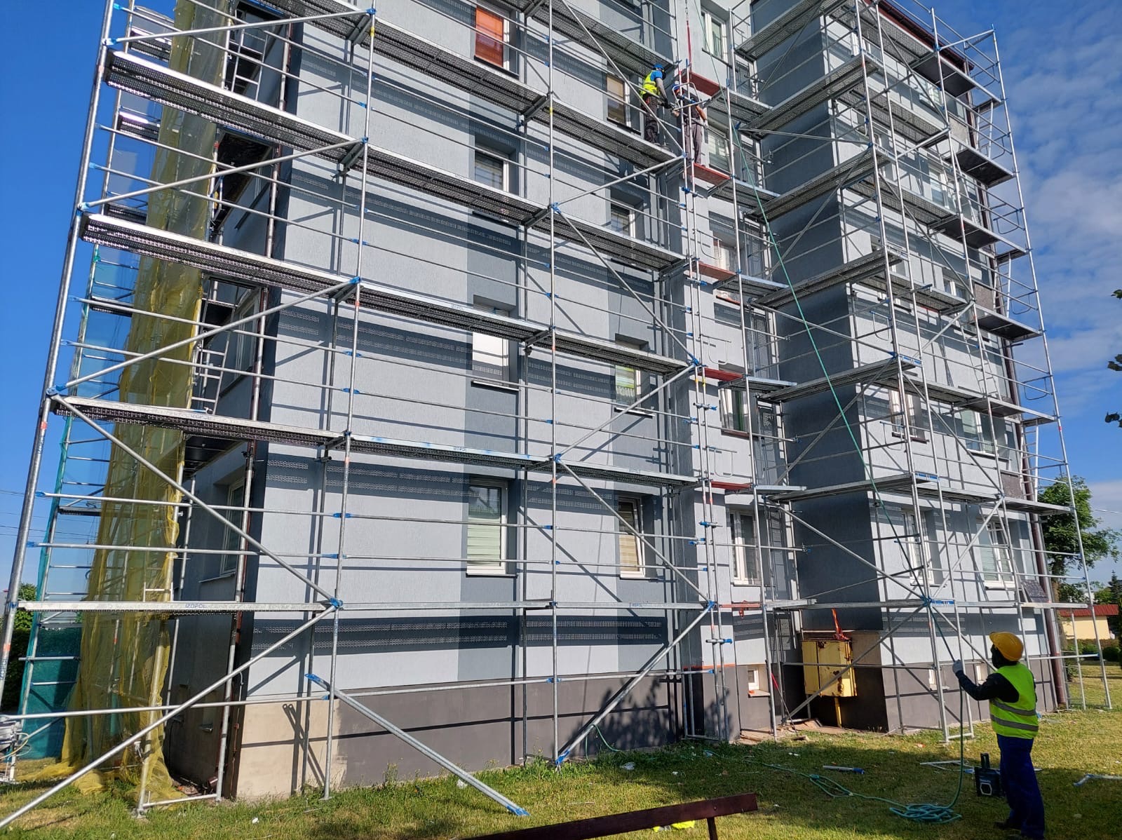 Szare, wielopiętrowe ocieplone rusztowanie elewacji budynku, z pracownikami na rusztowaniu i jednym na dole, w żółtej kamizelce, na tle błękitnego nieba.