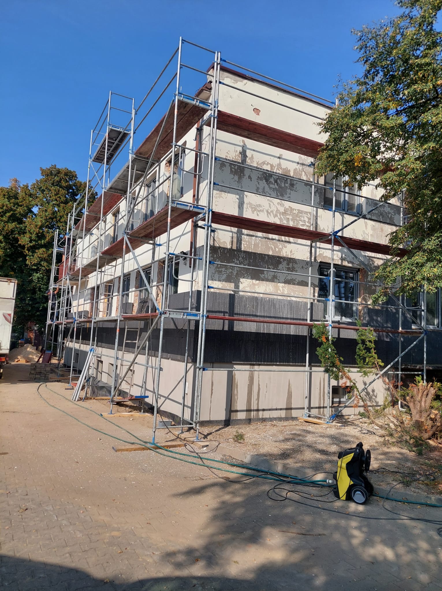 Budynek w trakcie ocieplania, z widocznymi rusztowaniami i fragmentami ocieplonej elewacji, na dole myjka ciśnieniowa. Częściowo ocieplona elewacja.