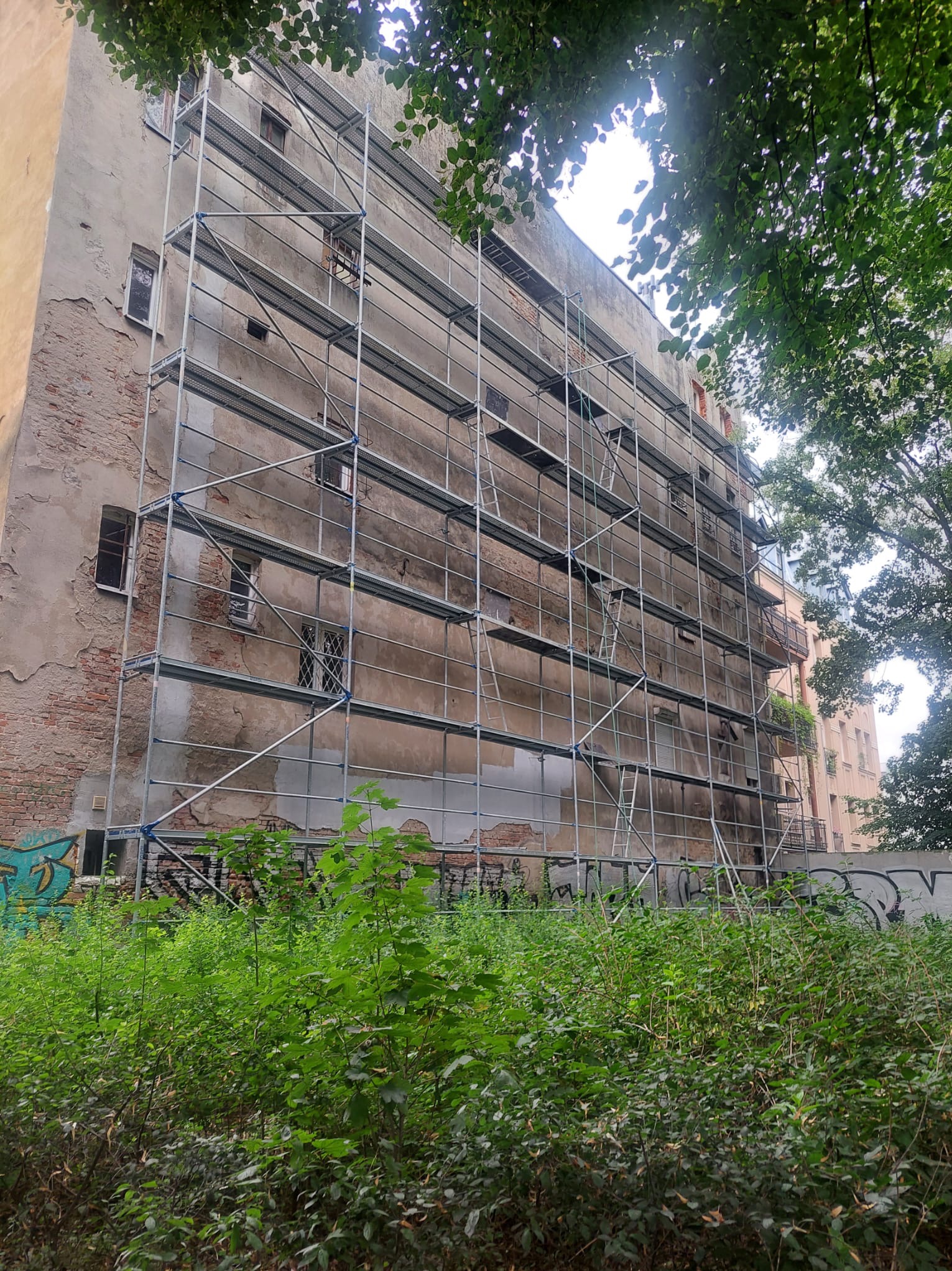 Elewacja budynku z widocznymi rusztowaniami, przygotowana do prac remontowych. Widoczne uszkodzenia tynku i fragmenty odsłoniętej cegły. Zieleń w dolnej części kadru.