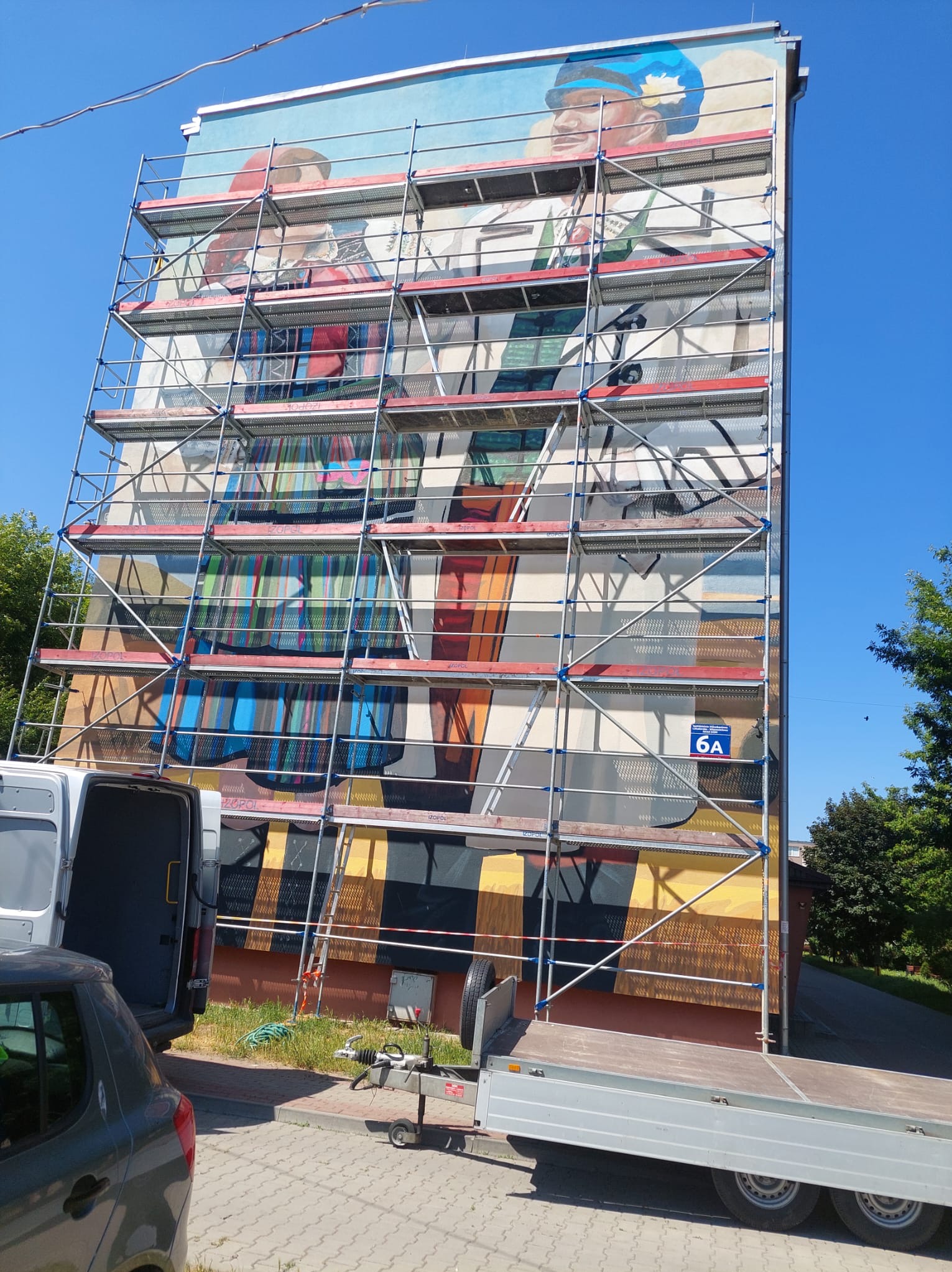 Elewacja budynku z muralem w trakcie renowacji, rusztowania na całej ścianie, widoczny fragment ulicy i zaparkowane pojazdy. Słoneczny dzień.