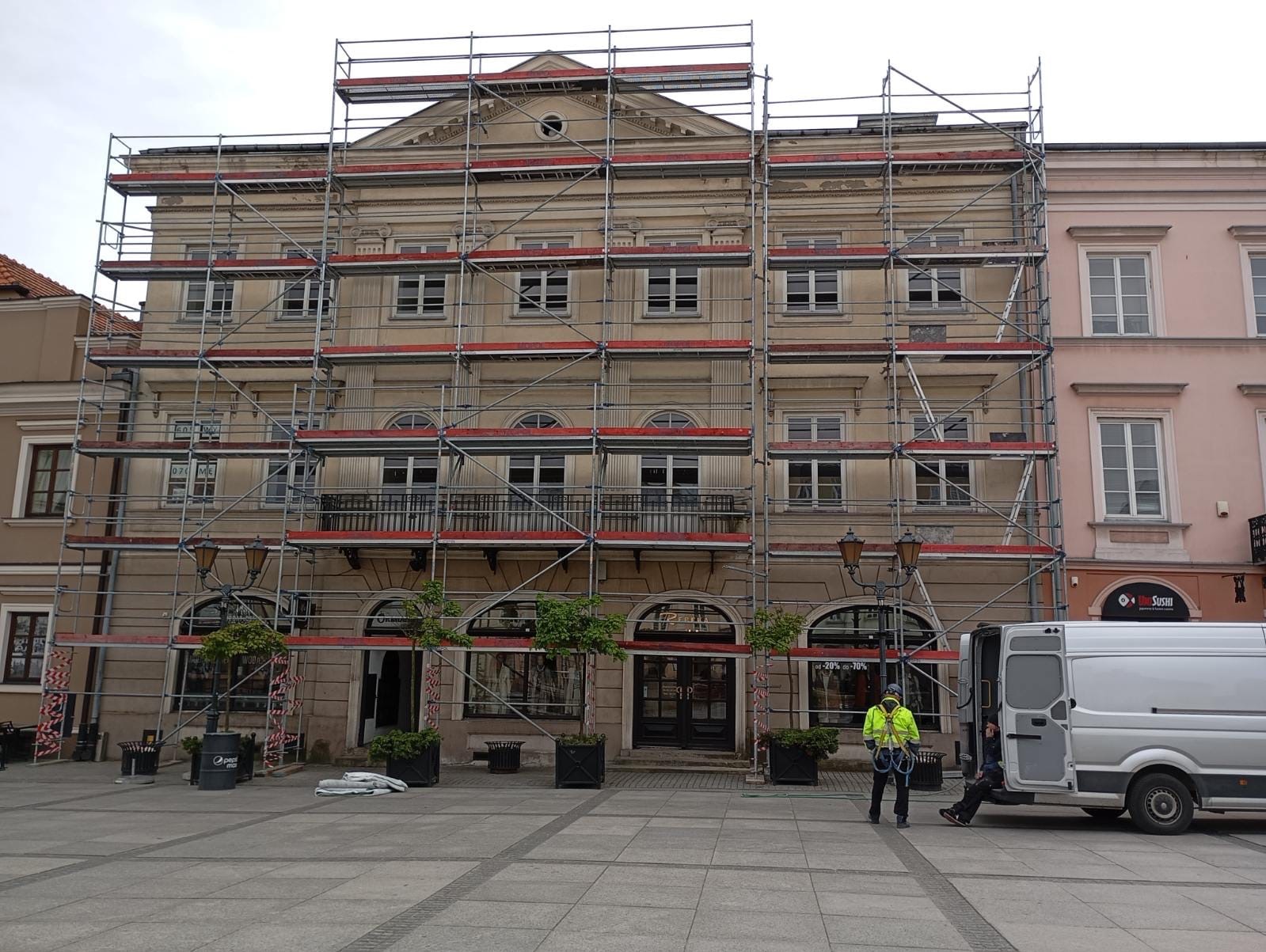 Elewacja kamienicy w rusztowaniach. Widoczny pracownik w odblaskowej kamizelce i biały bus. Prace remontowe na zewnątrz budynku w centrum miasta.