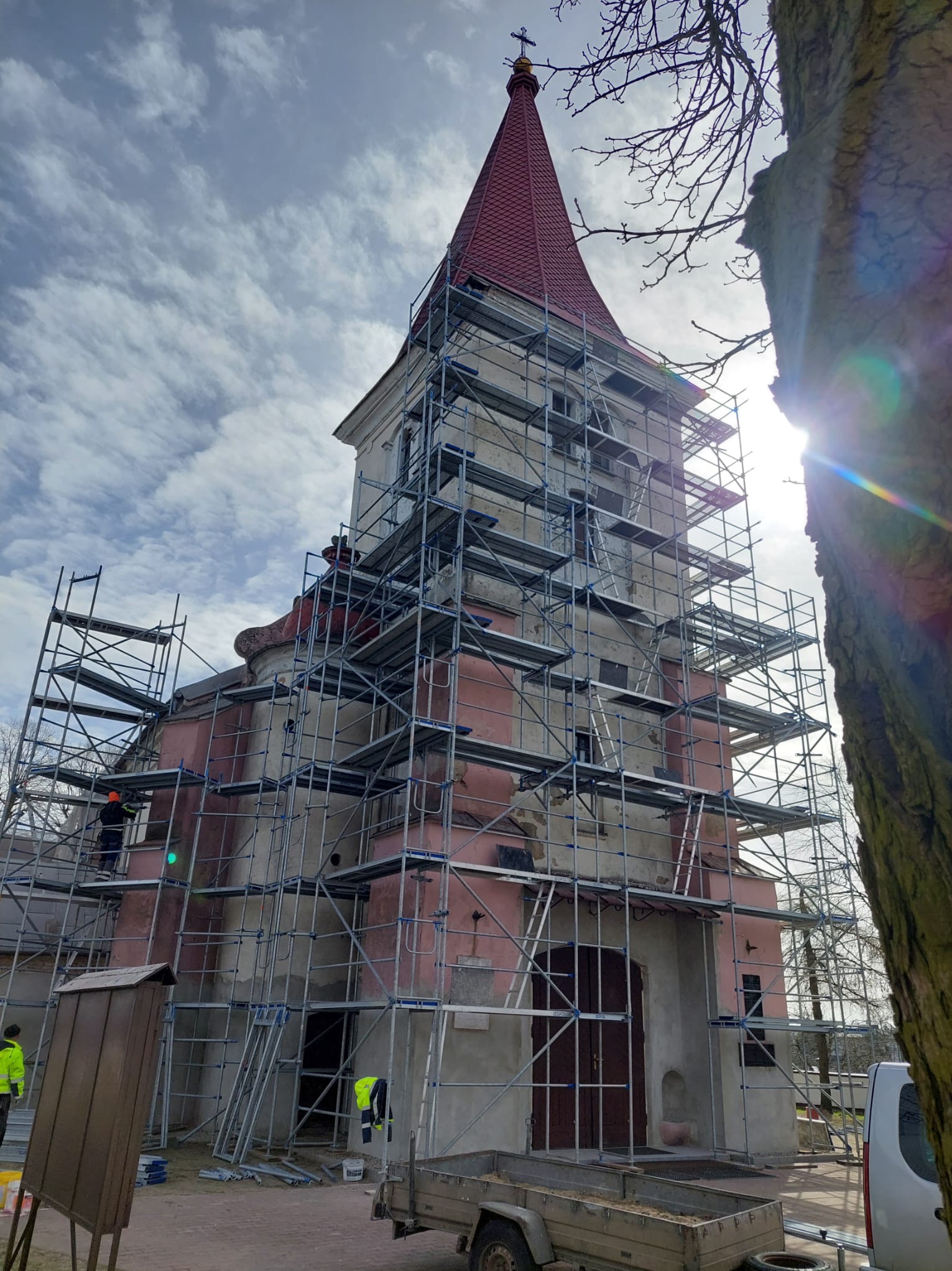 Remont elewacji kościoła z czerwoną wieżą.  Rusztowania okalają budynek, widoczni pracownicy w odblaskowych ubraniach. Słoneczny dzień, błękitne niebo z chmurami.