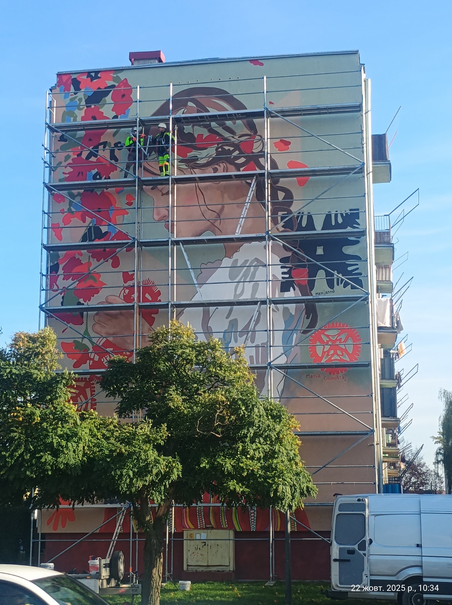 Wielkoformatowy mural z motywem kobiety i kwiatów na ścianie bloku, widoczny z poziomu ulicy, z rusztowaniem i pracownikami w trakcie realizacji.