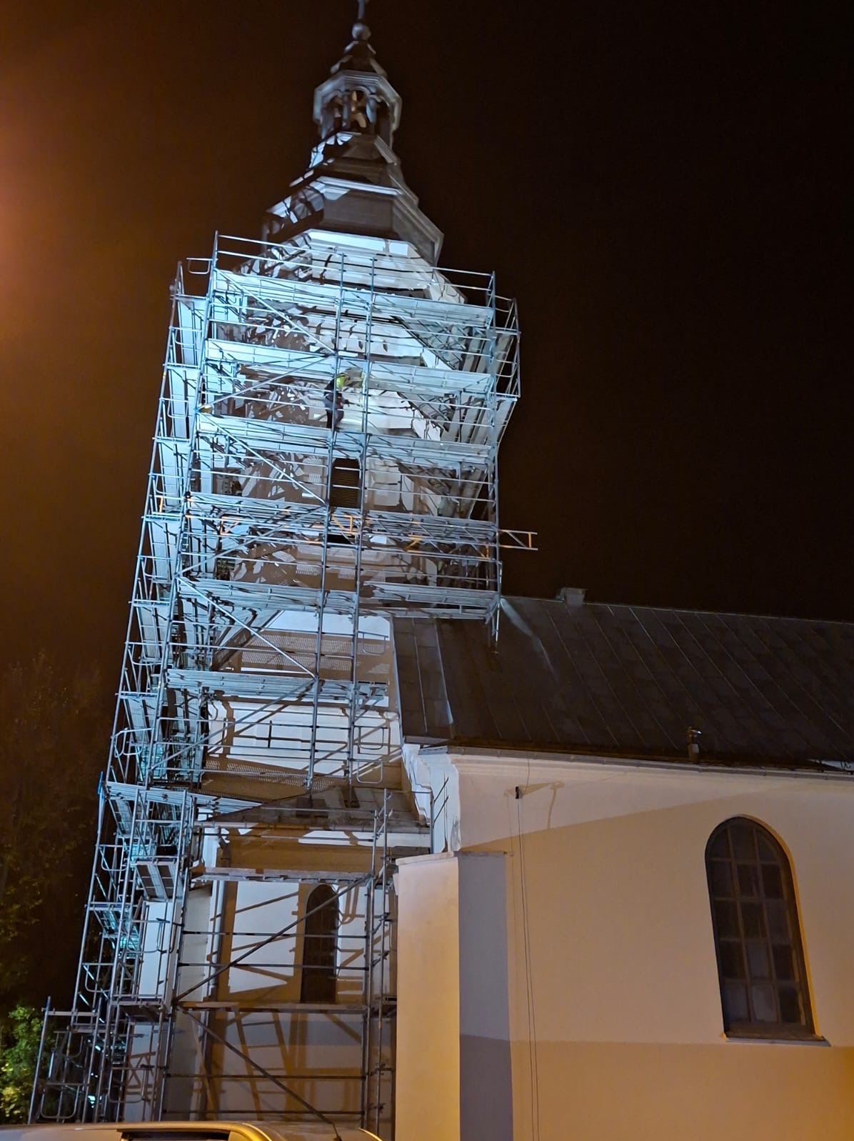 Nocne ujęcie wieży kościoła w rusztowaniach, oświetlonej sztucznym światłem. Widoczne fragmenty elewacji w trakcie prac remontowych.