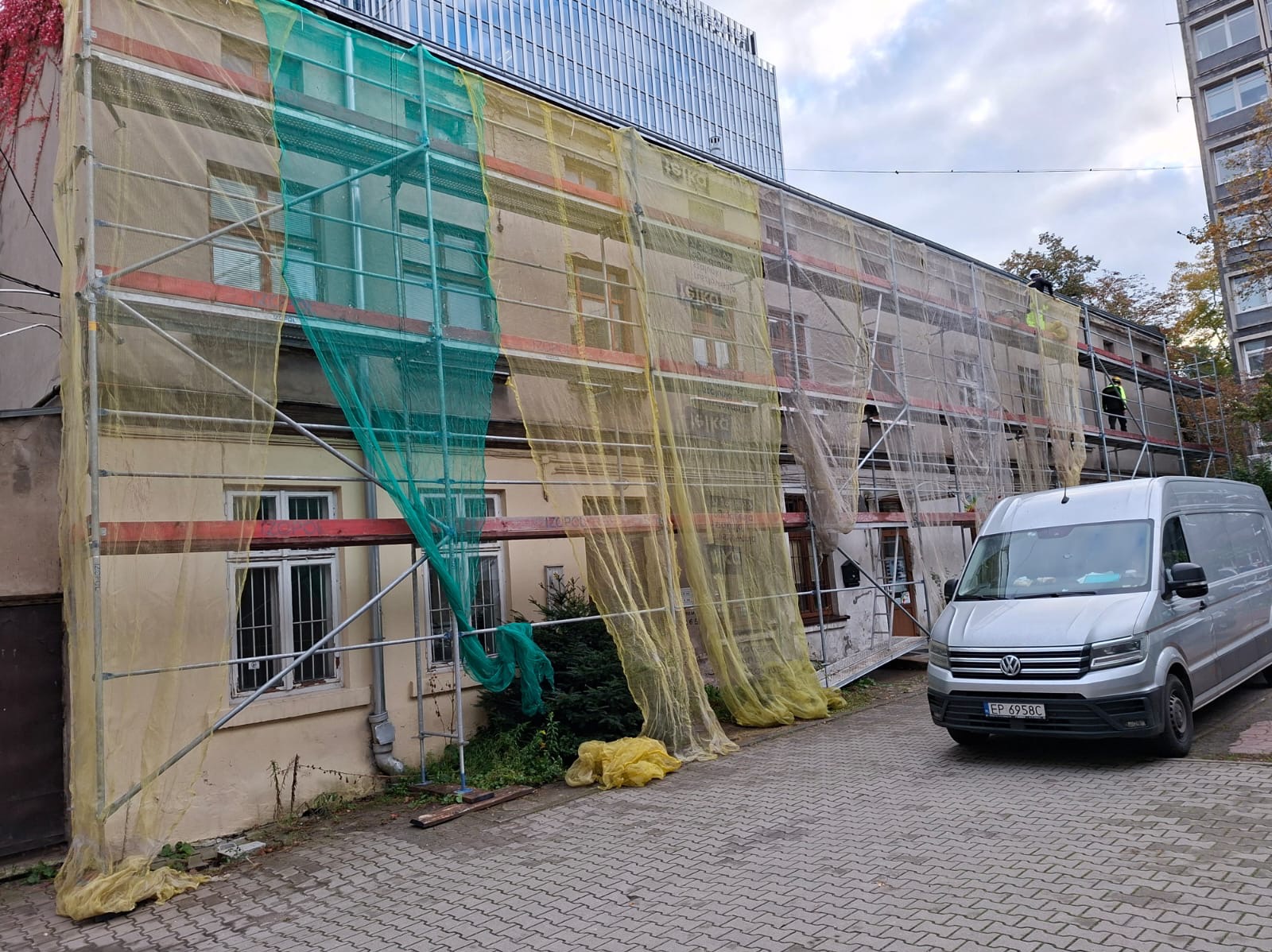 Budynek w trakcie ocieplania elewacji, osłonięty siatką ochronną na rusztowaniach. Na rusztowaniu pracownicy. Na pierwszym planie zaparkowany bus.