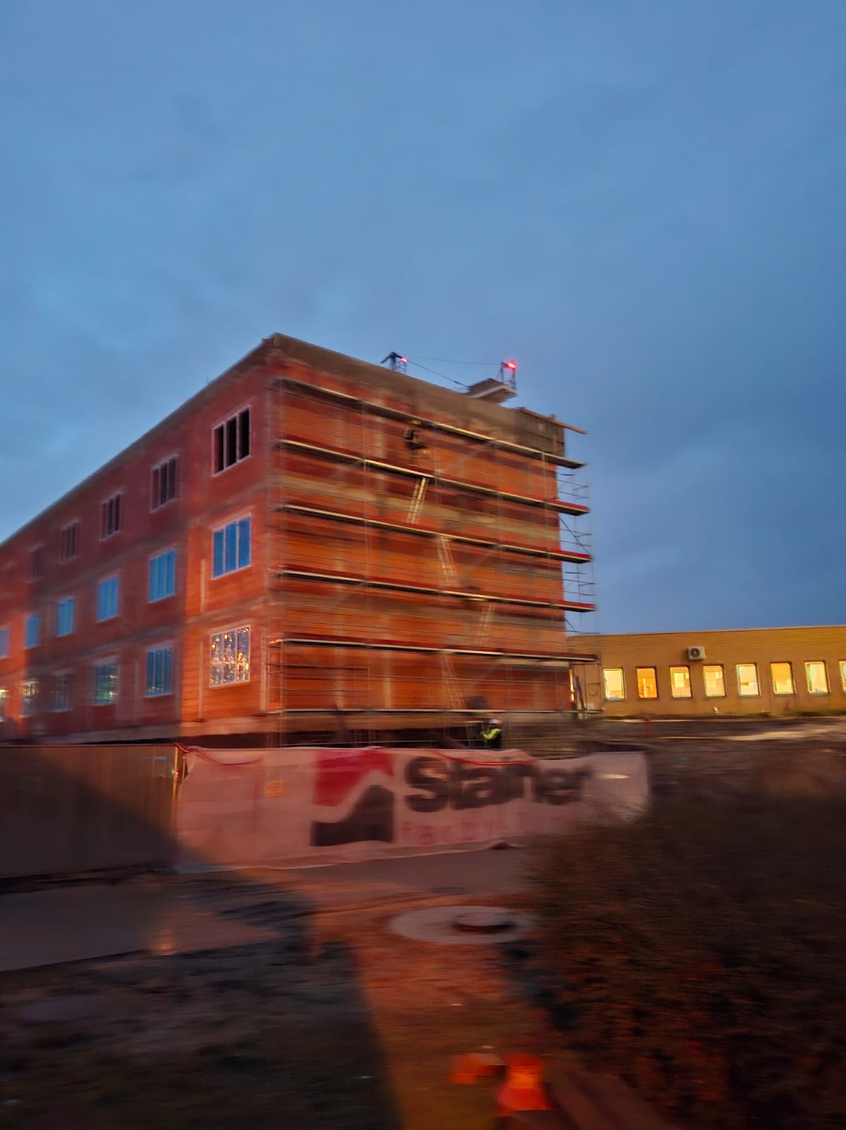 Budynek w trakcie budowy z cegły, otoczony rusztowaniami i siatką ochronną z logo. Widoczne okna z niebieskimi szybami i pracownik na rusztowaniu.