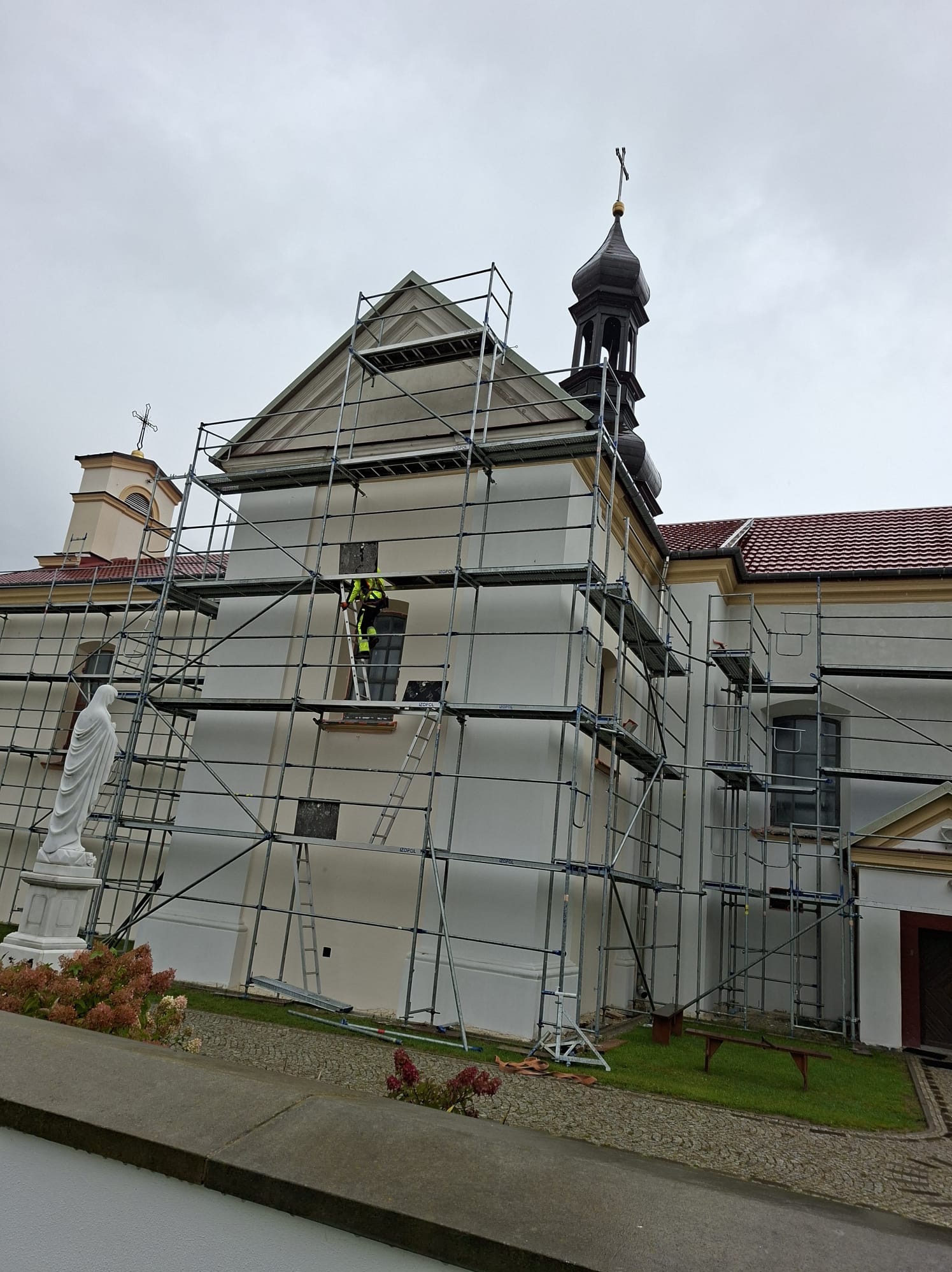 Prace na wysokościach przy kościele. Rusztowania obejmują budynek, a pracownik w odblaskowej kamizelce stoi na drabinie. W tle pochmurne niebo i statua.