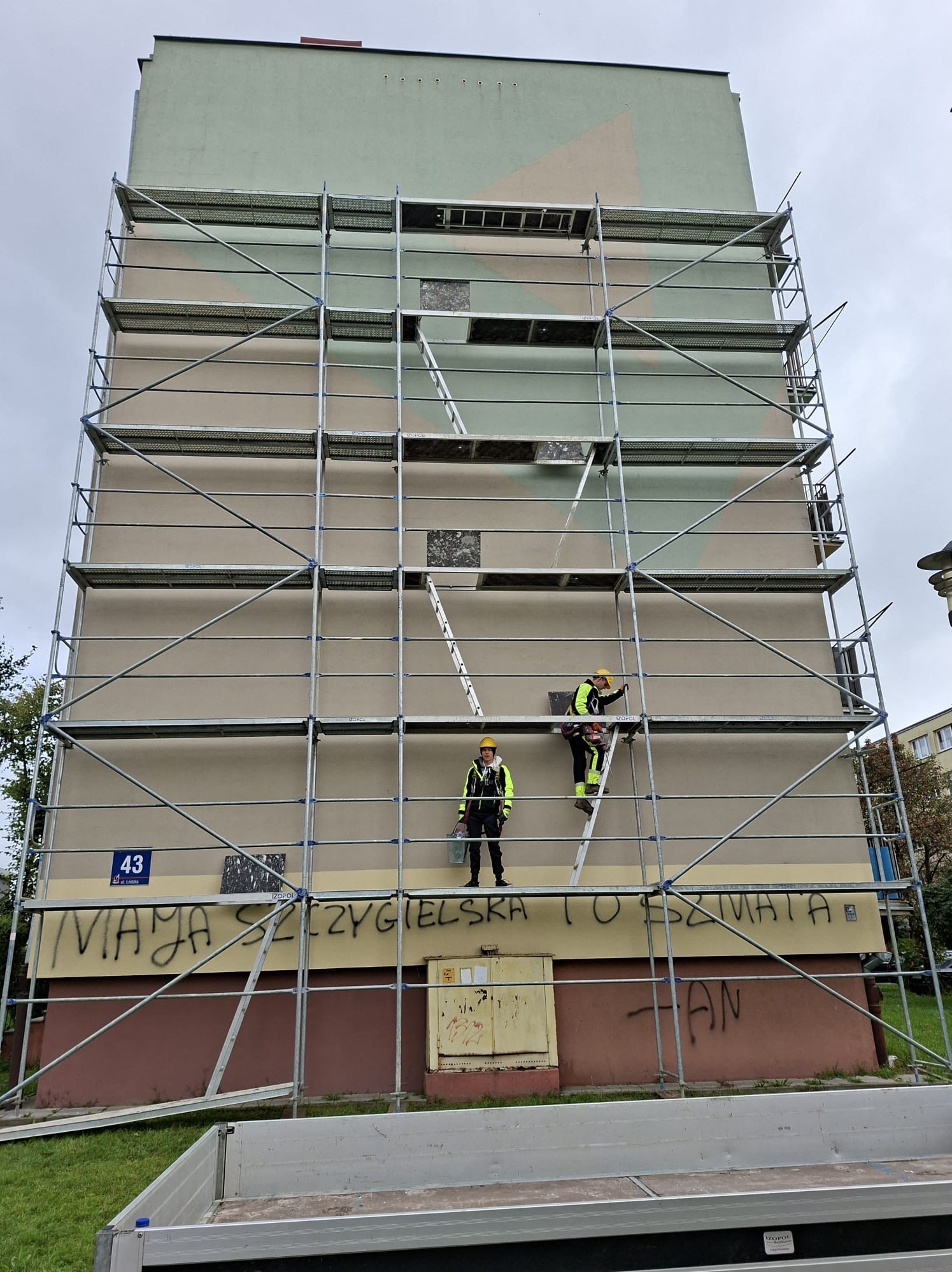 Pracownicy na rusztowaniu przy bloku z graffiti, przygotowujący elewację do ocieplenia. Widoczne fragmenty ocieplenia i drabina. Ujęcie z dołu, pochmurne niebo.