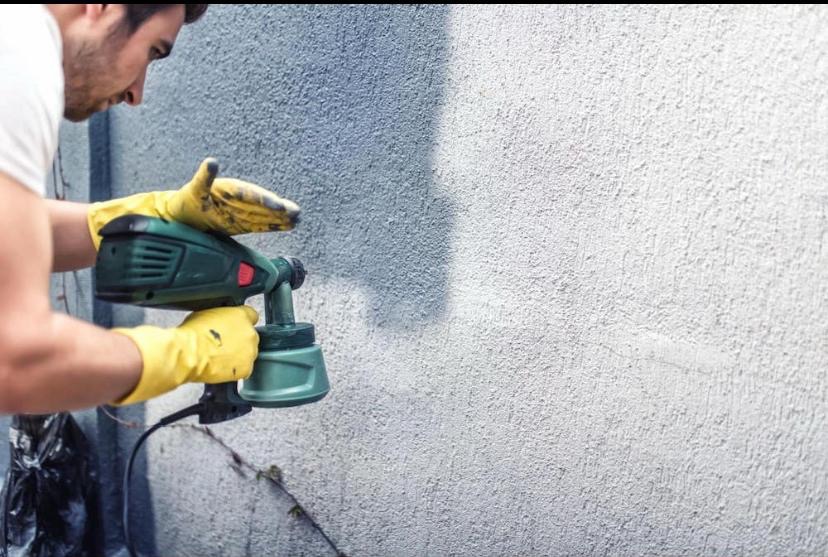 Malowanie elewacji natryskowo: mężczyzna w rękawicach aplikuje farbę na szorstką ścianę za pomocą pistoletu natryskowego. Widoczne świeżo pomalowane fragmenty.