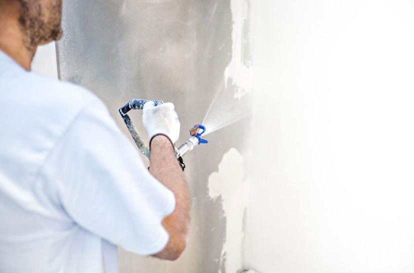 Pracownik w białej koszulce natryskuje białą farbę na ścianę za pomocą pistoletu natryskowego. Widoczne świeże ślady farby na szarej ścianie.