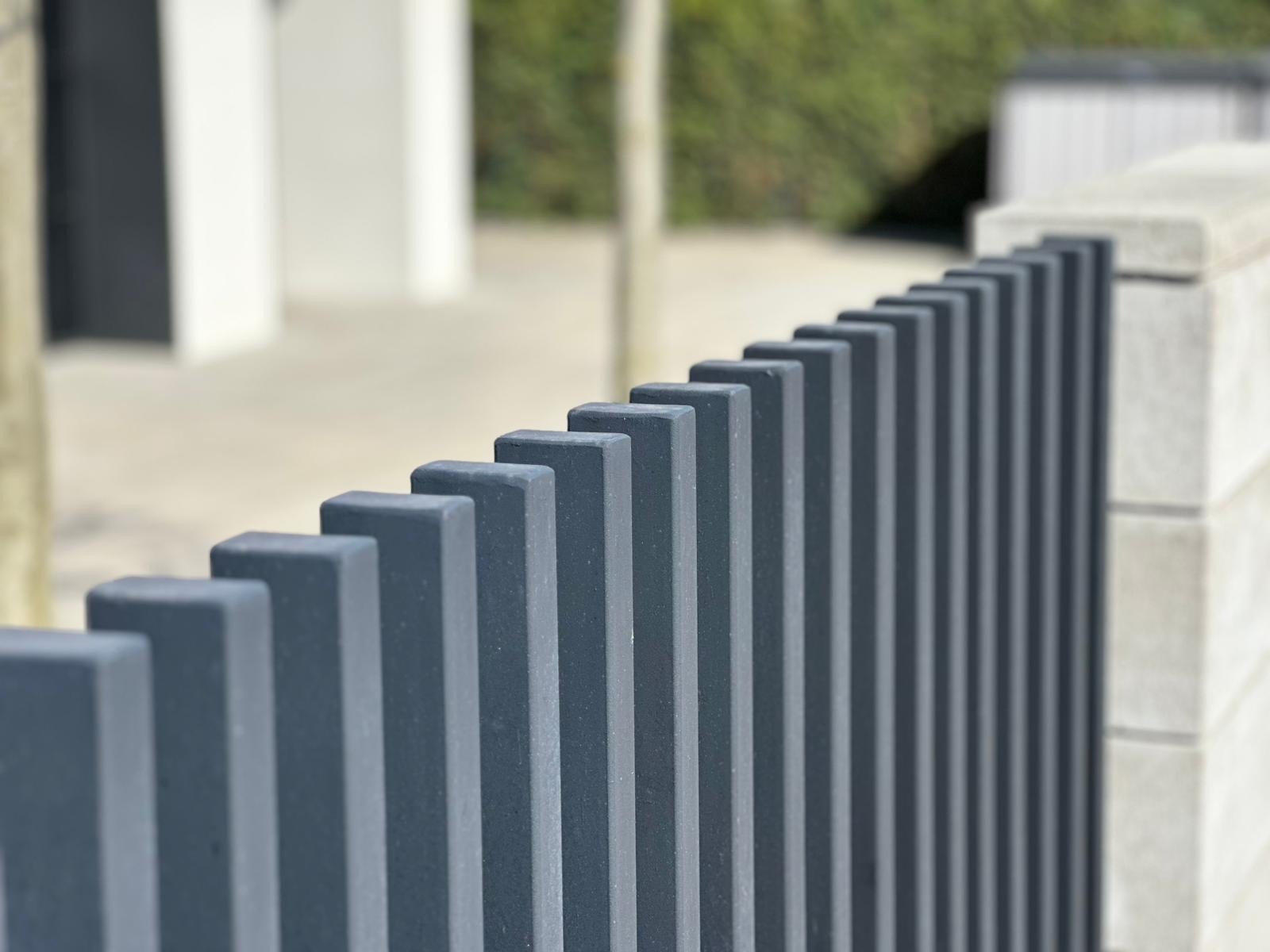 Nowoczesne, grafitowe ogrodzenie panelowe z betonu architektonicznego, widziane pod kątem, z rozmytym tłem zieleni i jasnej elewacji budynku.
