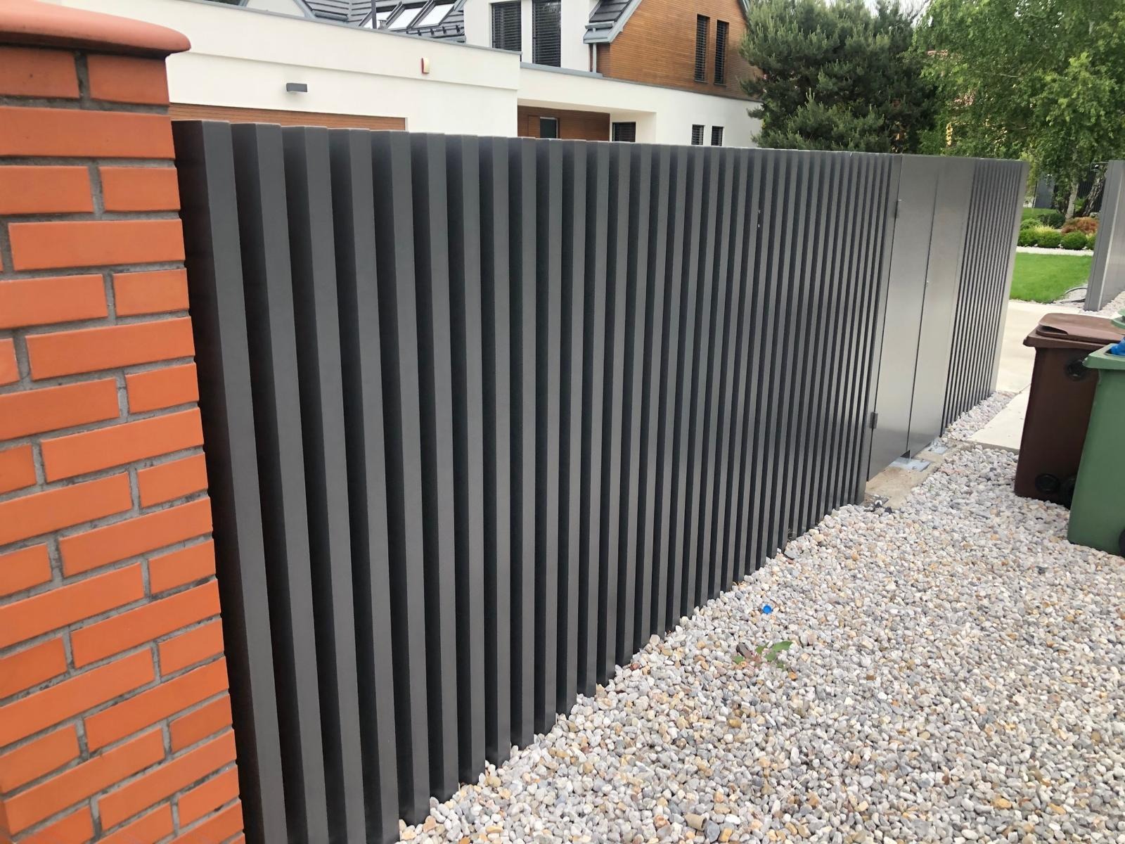 Nowoczesne ogrodzenie z pionowych, grafitowych paneli, sąsiadujące z ceglanym słupkiem. Białe kamienie na ziemi, w tle dom i zieleń.