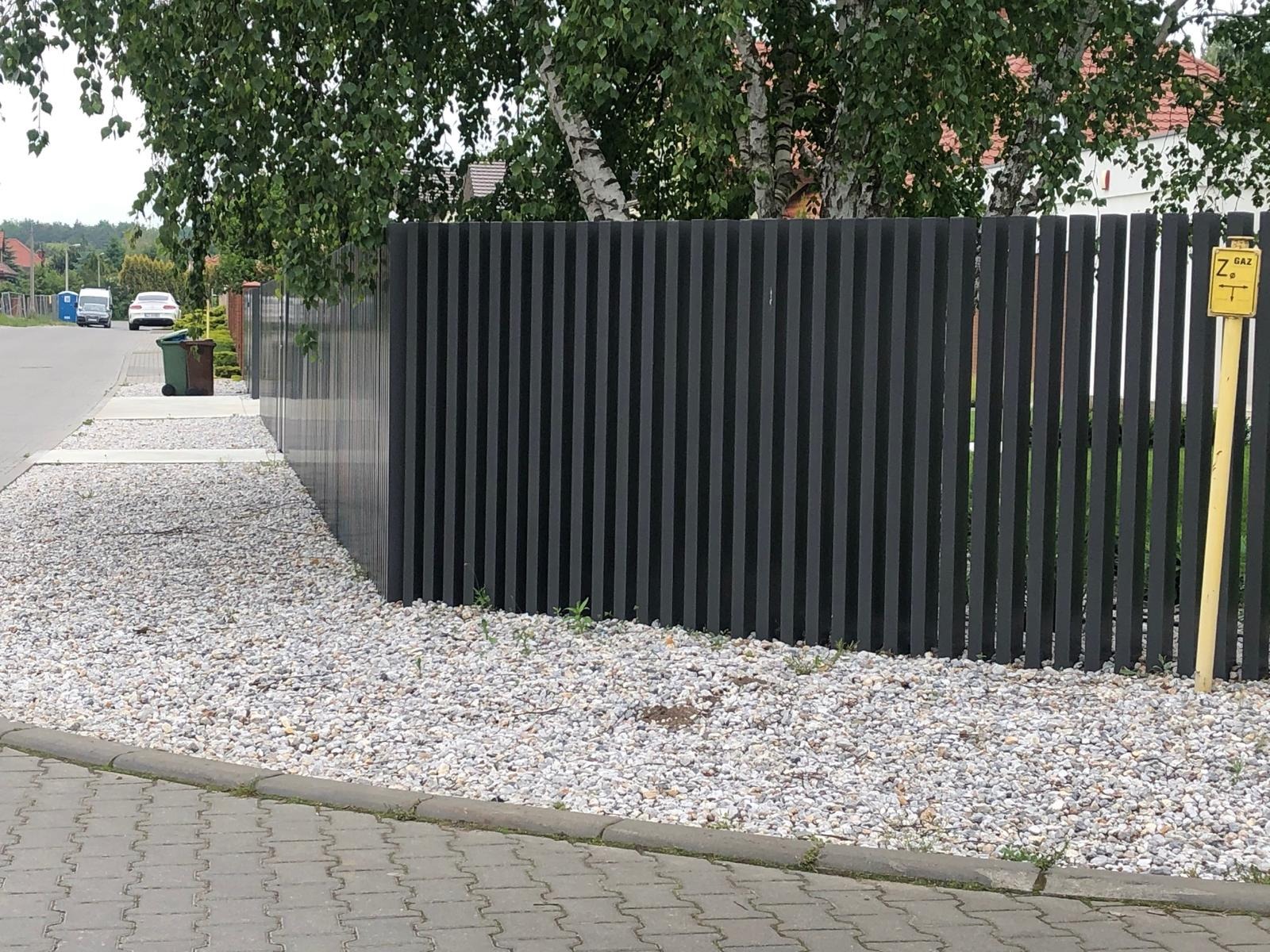 Nowoczesne ogrodzenie panelowe w kolorze grafitowym, otoczone białym żwirem, z widocznym znakiem gazowym i fragmentem chodnika z kostki brukowej.
