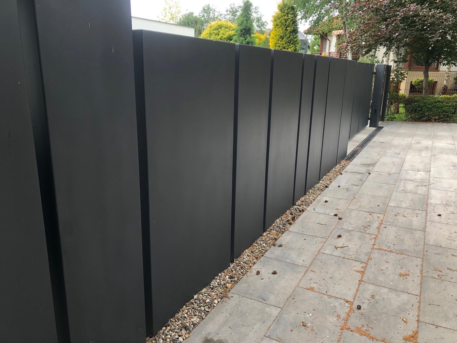 Nowoczesne, grafitowe ogrodzenie panelowe z wąskimi, pionowymi przerwami, przylegające do wyłożonego kostką brukową podjazdu z drobnym żwirem.