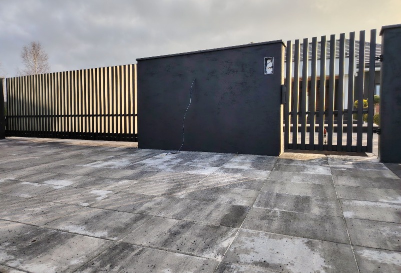 Nowoczesna, grafitowa brama posesji z pionowymi szczeblami, wykończona kostką brukową. Widoczne elementy instalacji elektrycznej na ścianie bramy.