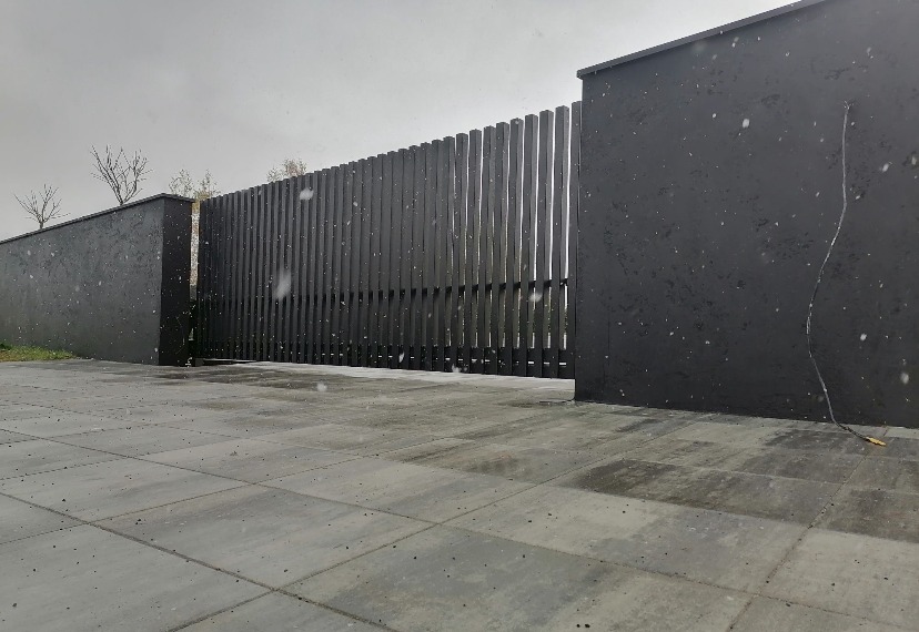 Nowoczesna, grafitowa brama posesyjna z pionowych desek, wkomponowana w mur z elementami roślinności. Kostka brukowa na pierwszym planie, pochmurne niebo w tle.