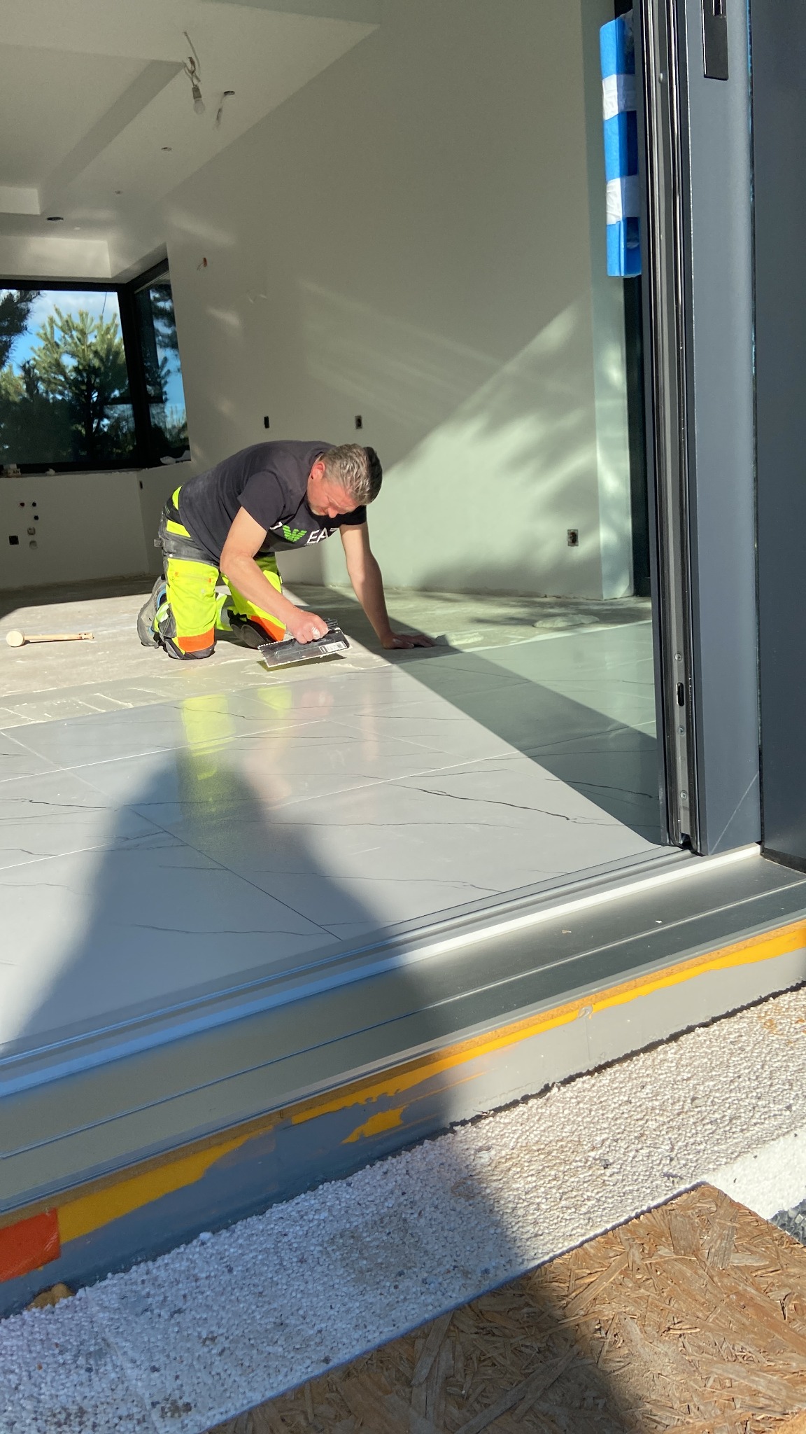 Glazurnik układa szare płytki podłogowe w jasnym wnętrzu. Widoczny fragment okna i drzwi przesuwnych. Mężczyzna w odzieży roboczej na kolanach, używa kielni z zębami.