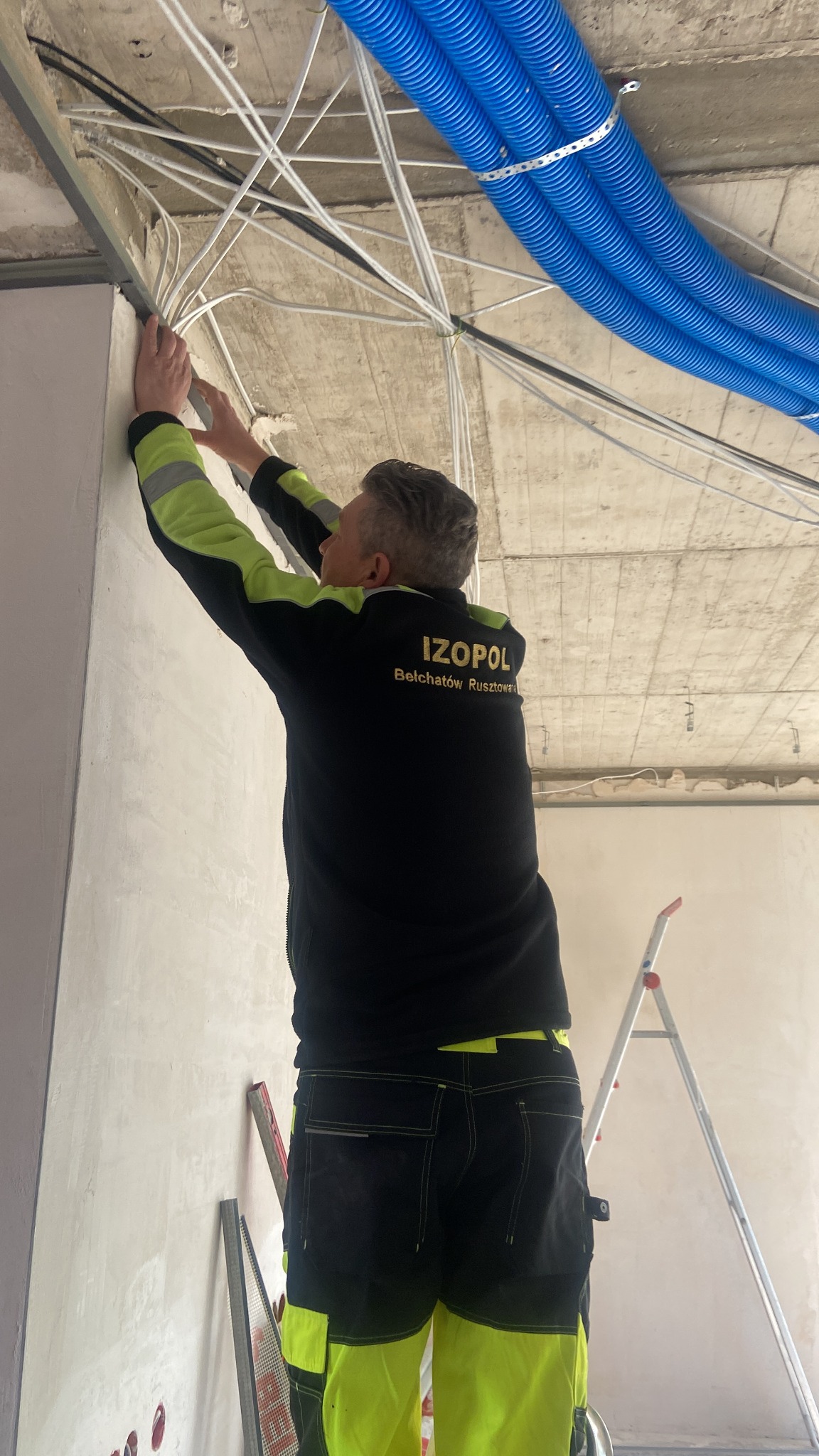 Pracownik w firmowej bluzie montuje instalacje pod sufitem. Widoczne niebieskie rury i białe przewody. W tle drabina i fragment ściany. Ujęcie od dołu.