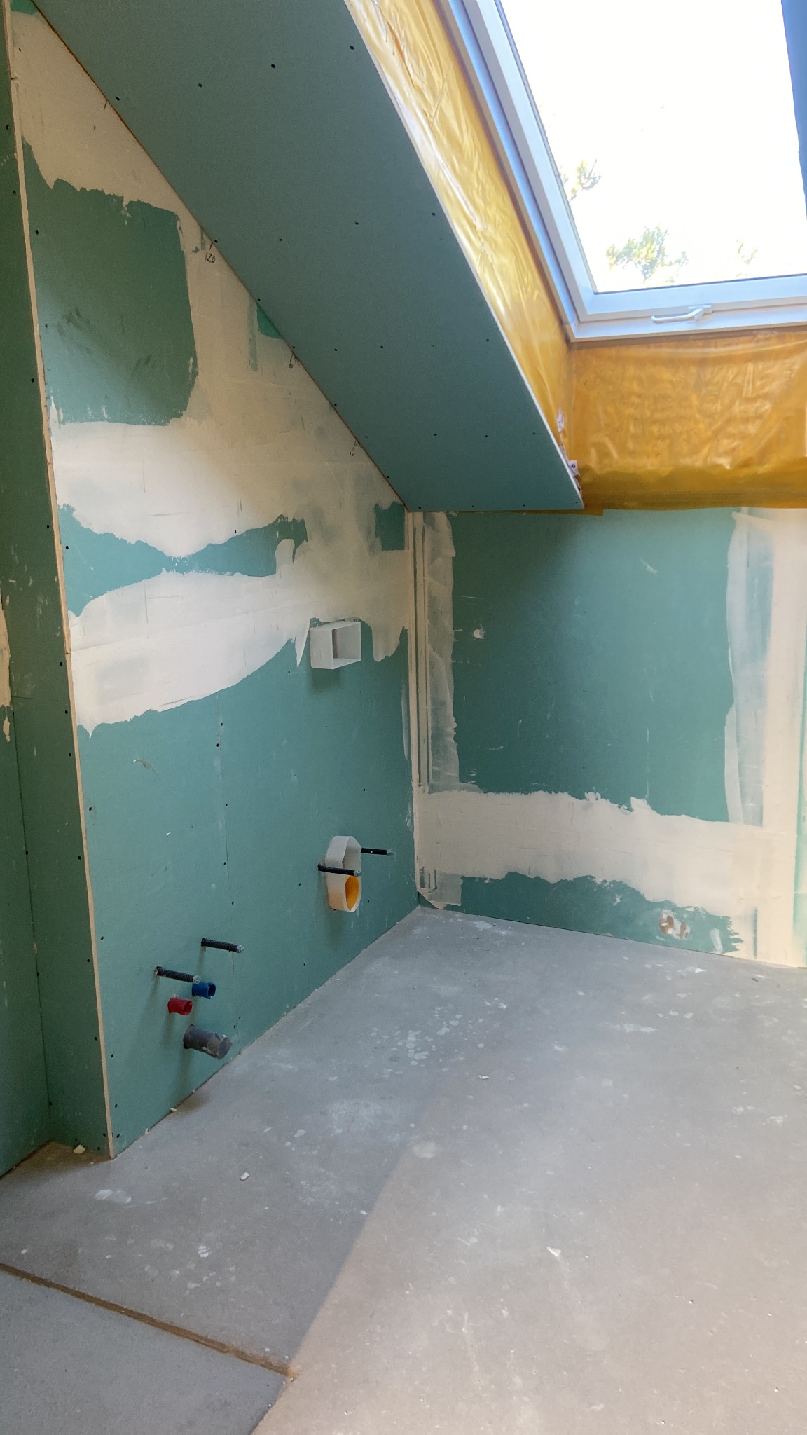 Surowe wnętrze z płytami gipsowo-kartonowymi, widoczne instalacje hydrauliczne i okno dachowe. Ściany przygotowane do dalszych prac wykończeniowych.