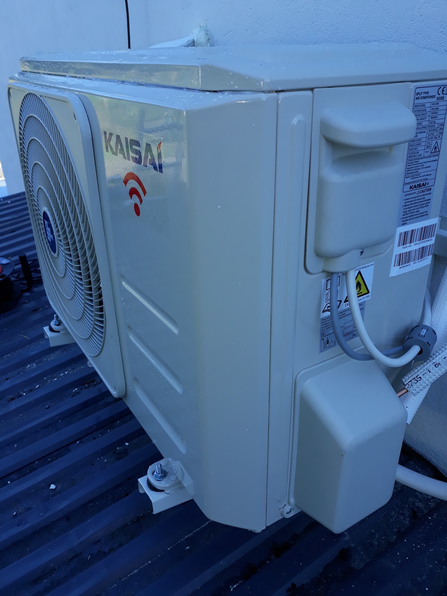 Zewnętrzna jednostka klimatyzacji Kaisaia z widocznym logo i oznaczeniem WiFi, zamontowana na dachu z blachy trapezowej, widoczne przewody i naklejki informacyjne.