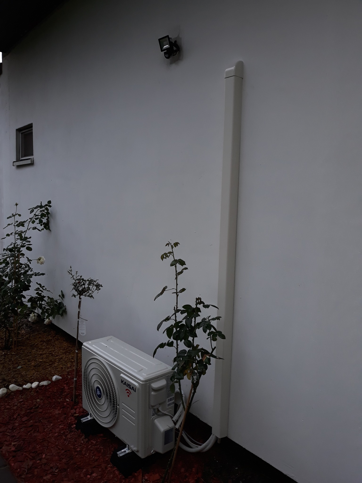 Zewnętrzna jednostka klimatyzacji Kaisai zamontowana przy białej ścianie domu, otoczona rabatą z korą i młodymi krzewami róż.