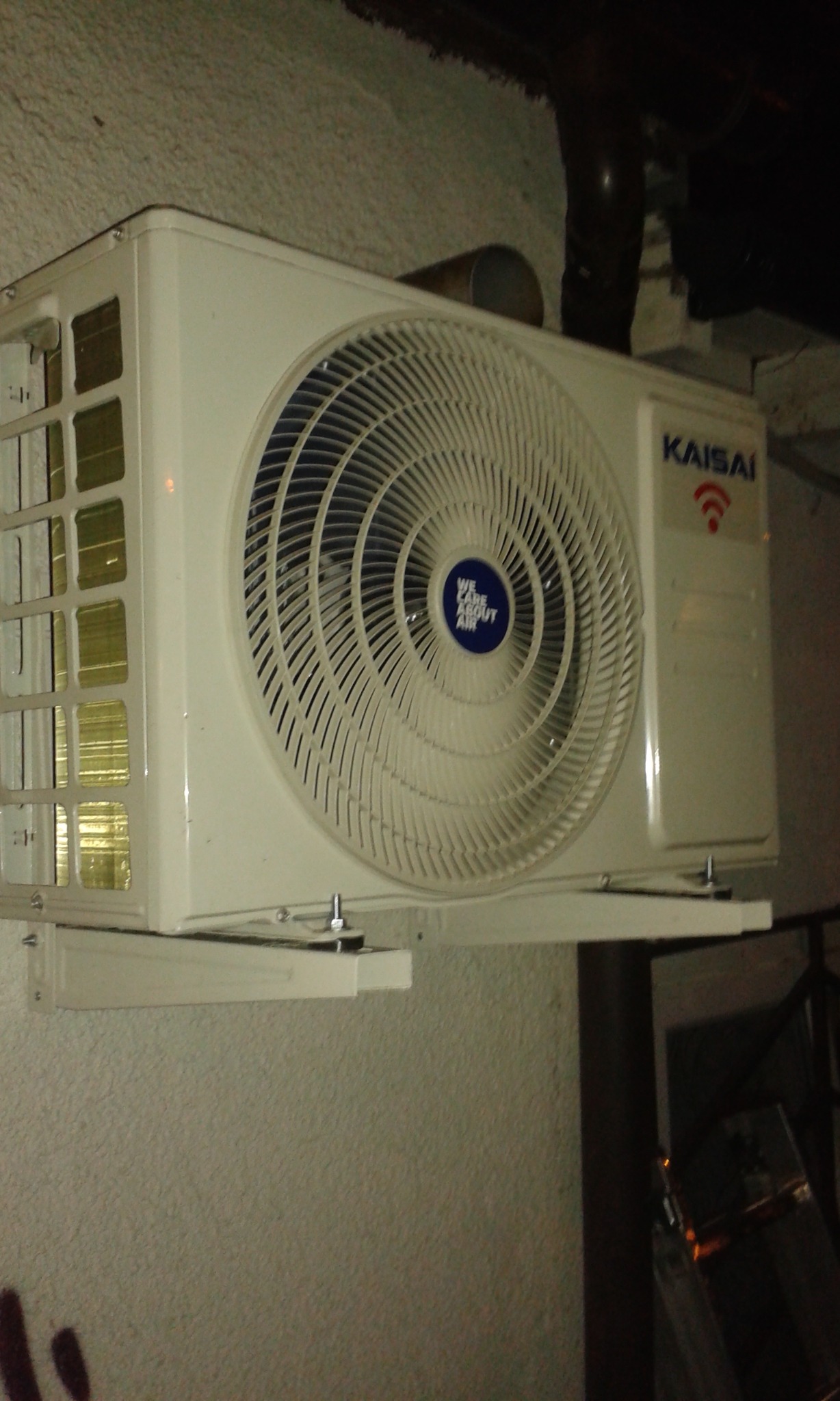 Biała jednostka zewnętrzna klimatyzacji Kaisai zamontowana na ścianie z widocznymi śrubami montażowymi i rurą odprowadzającą, sfotografowana z dołu w słabym oświetleniu.