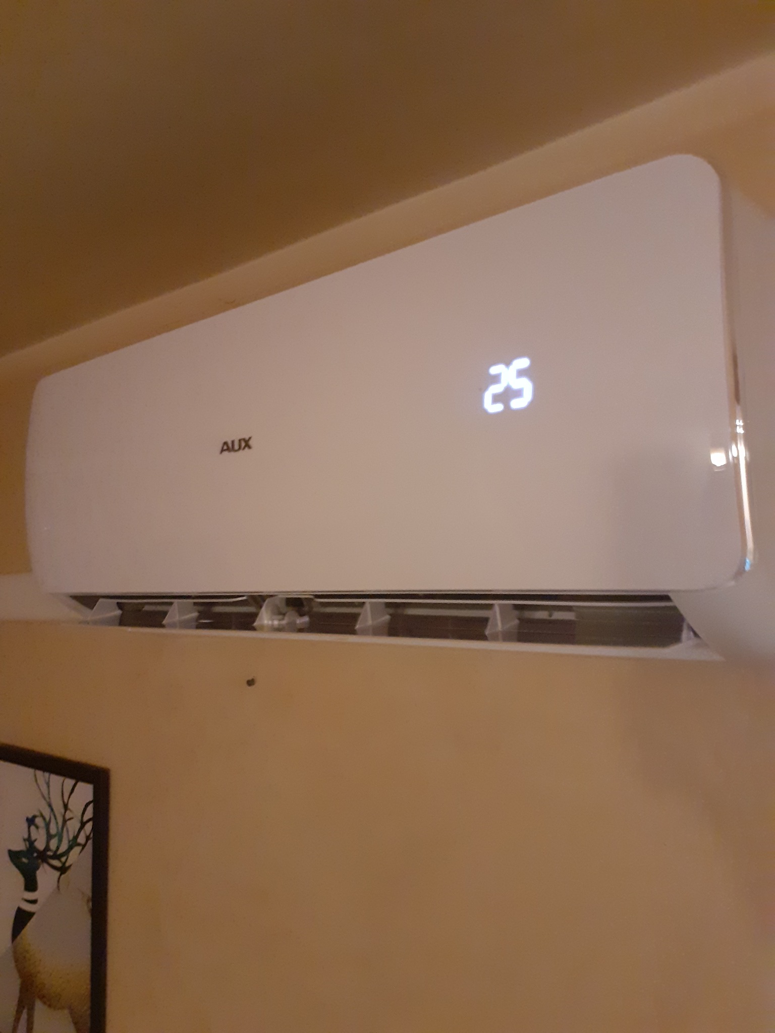 Biała klimatyzacja marki AUX zamontowana na beżowej ścianie, wyświetlająca temperaturę 25 stopni Celsjusza, w tle widoczny obraz z motywem jelenia.