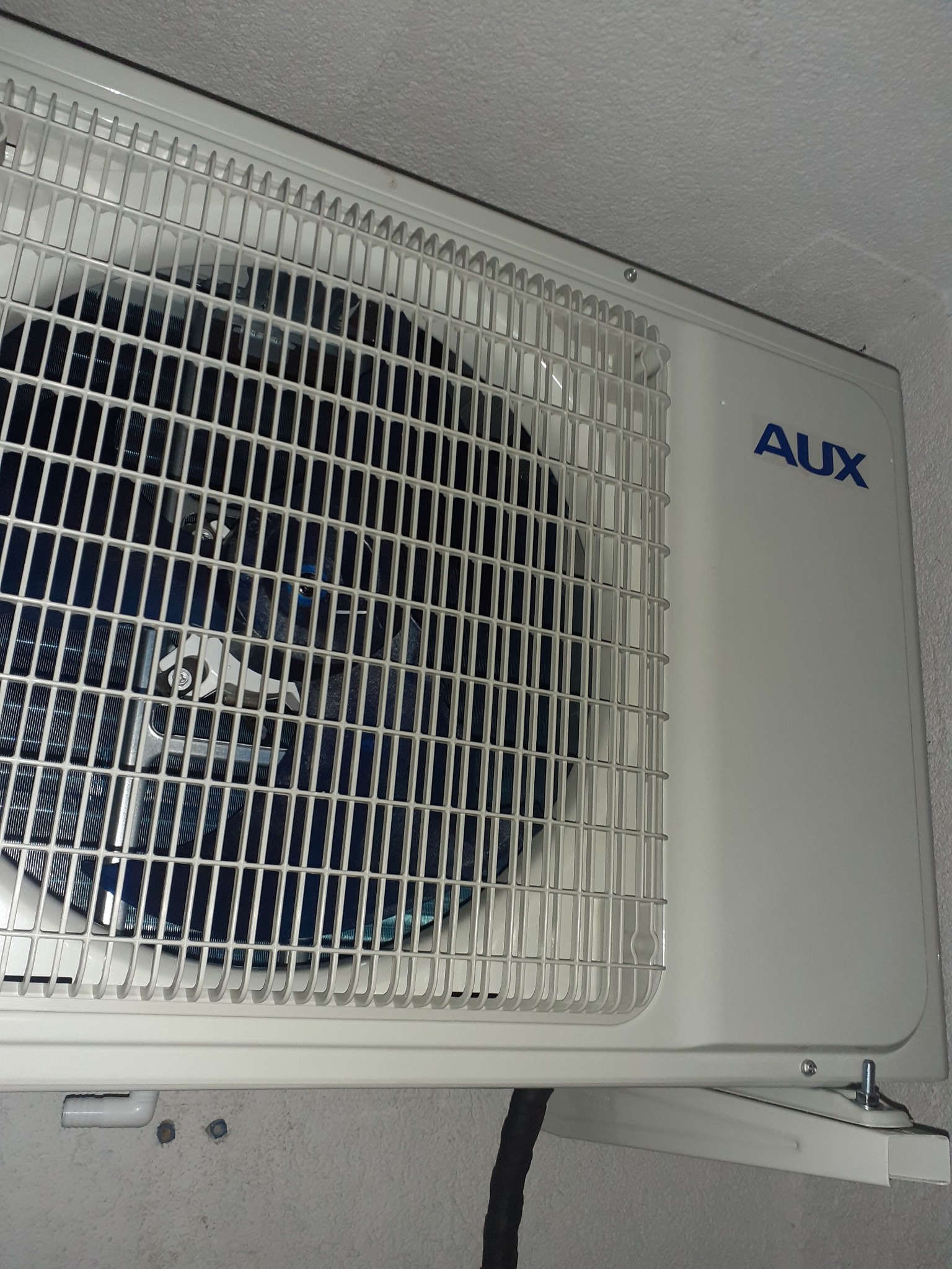 Biała jednostka wewnętrzna klimatyzacji AUX zamontowana na szarej ścianie, widoczna kratka wlotu powietrza i niebieskie logo marki.