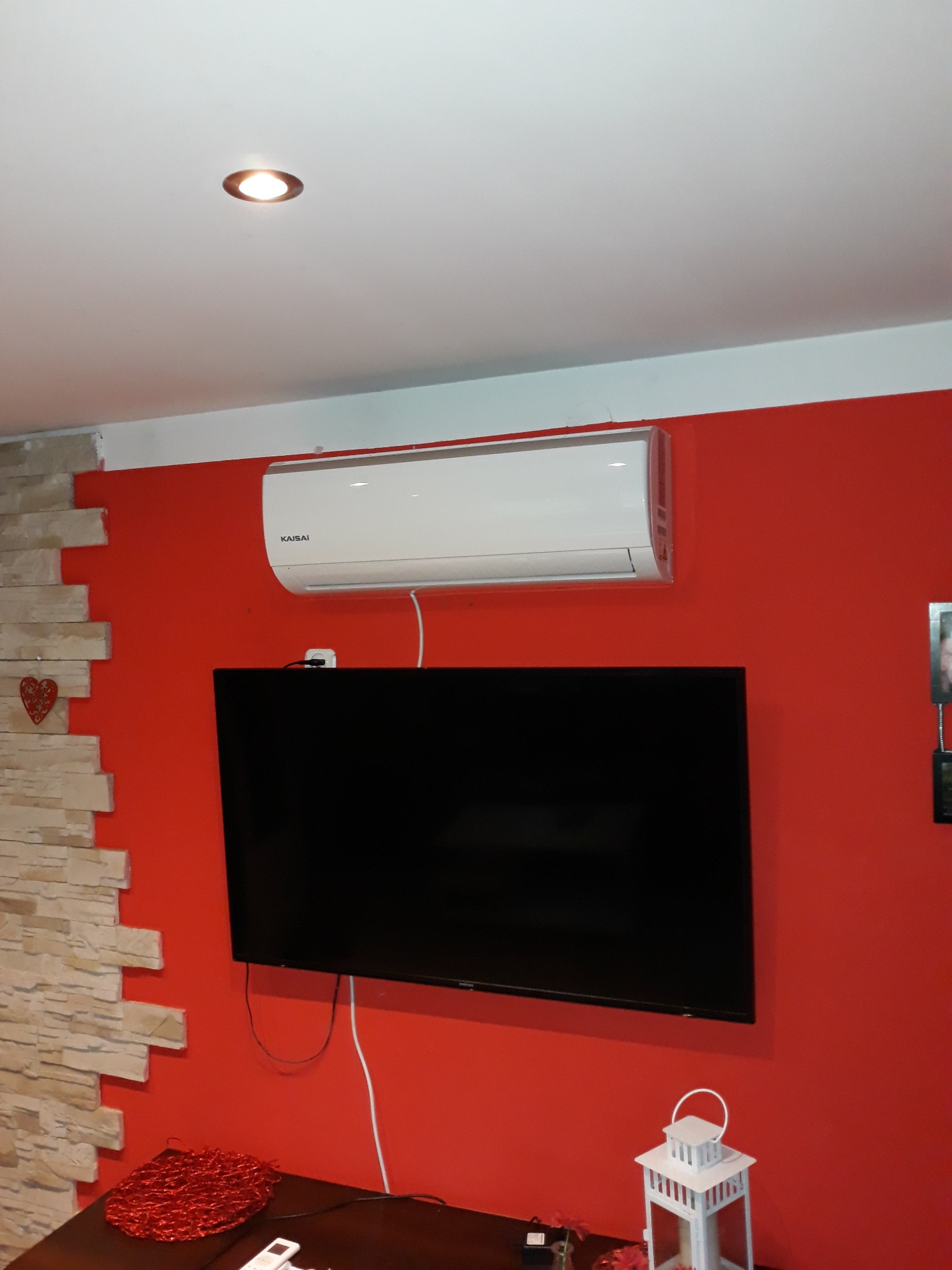 Biała klimatyzacja Kaisai zamontowana nad czarnym telewizorem na czerwonej ścianie w salonie z dekoracyjnym kamieniem i lampką nocną.