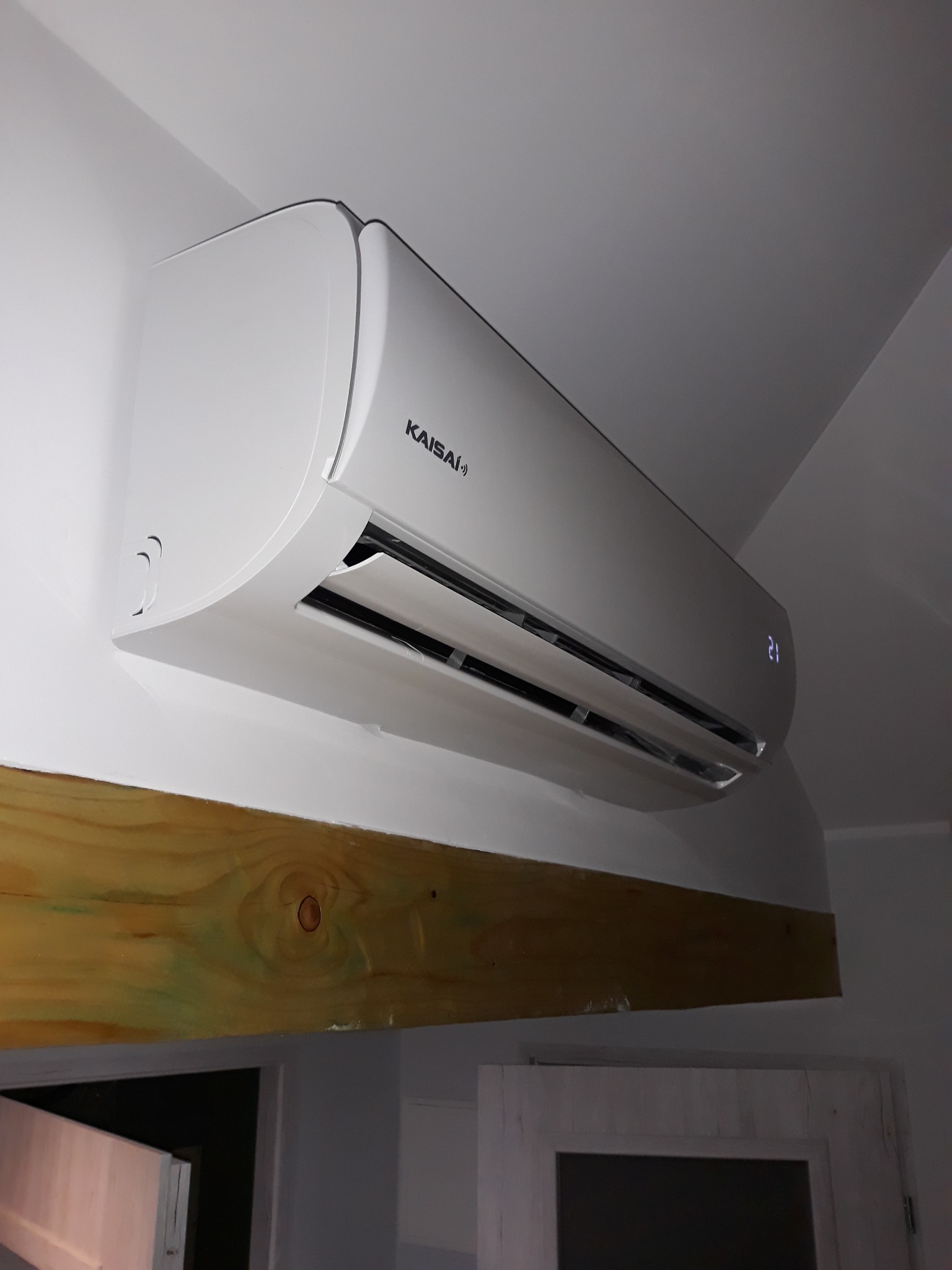 Biała jednostka klimatyzacji Kaisai zamontowana pod sufitem, widoczna drewniana belka stropowa i fragment białych drzwi.