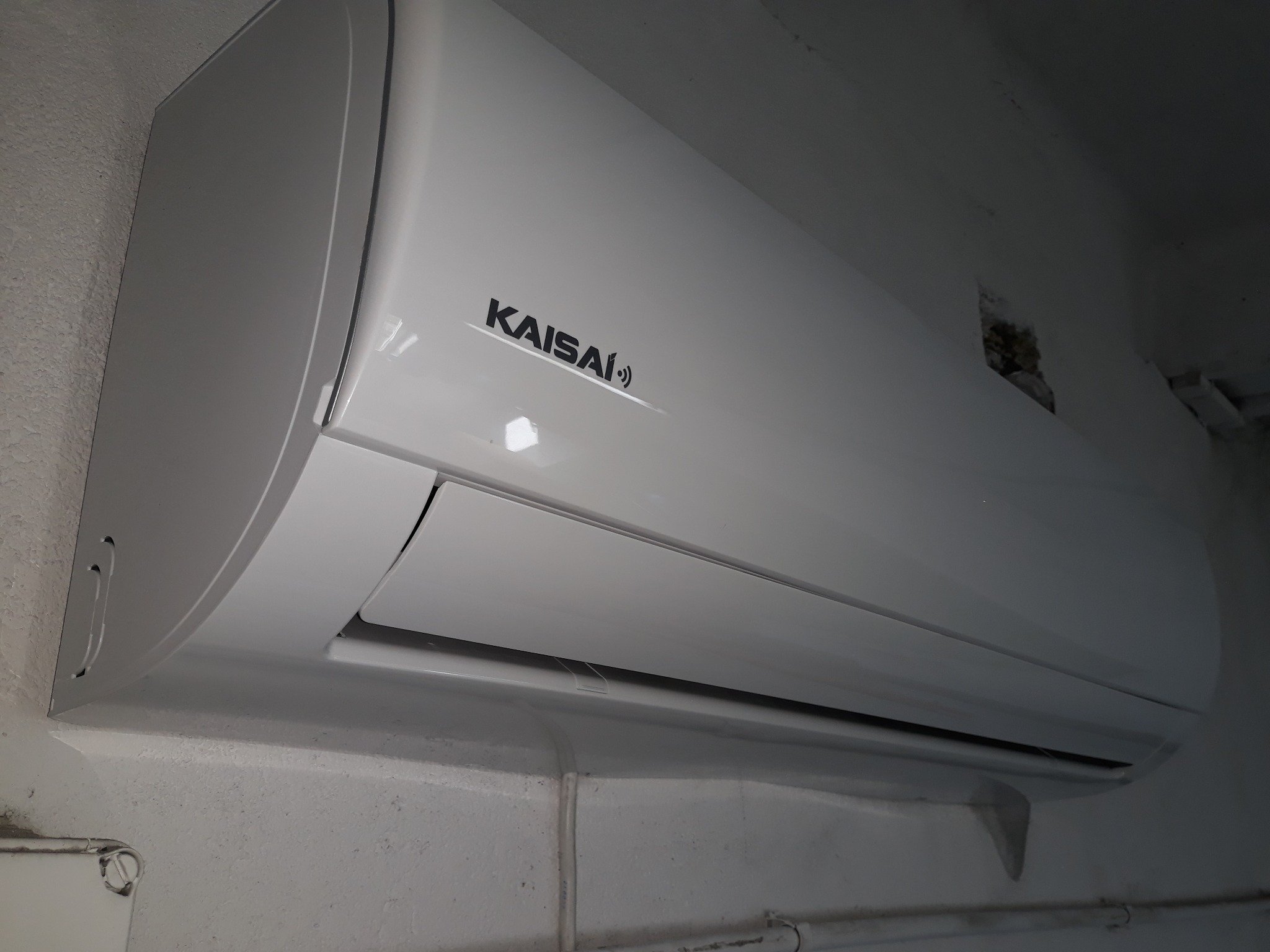 Biała jednostka wewnętrzna klimatyzacji Kaisai zamontowana na pomalowanej ścianie z widocznymi instalacjami elektrycznymi w listwach.