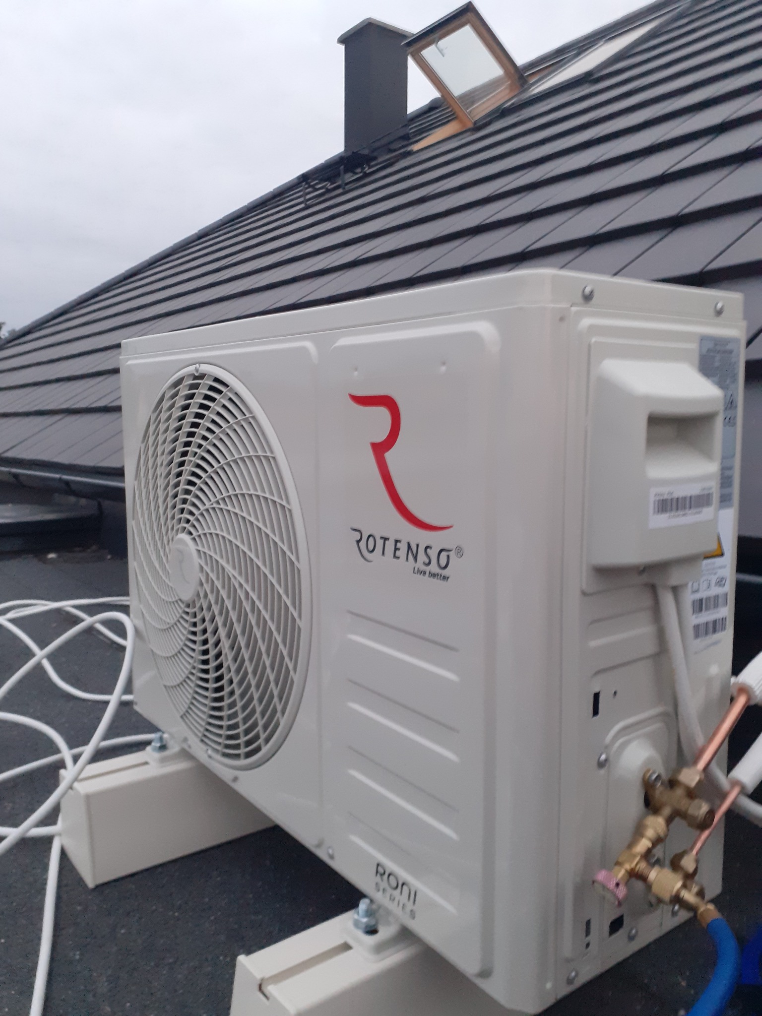Zewnętrzna jednostka klimatyzacji Rotenso zamontowana na dachu domu z szarą dachówką, widoczne miedziane rurki i białe kable.