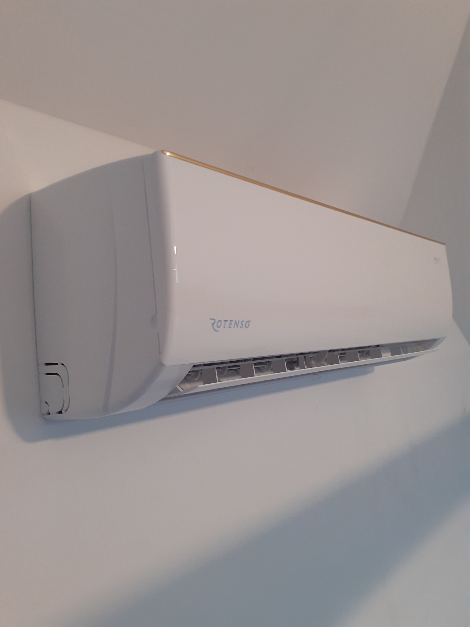 Biała jednostka klimatyzacji Rotenso, zamontowana na jasnej ścianie, z delikatnym złotym akcentem na górnej krawędzi.