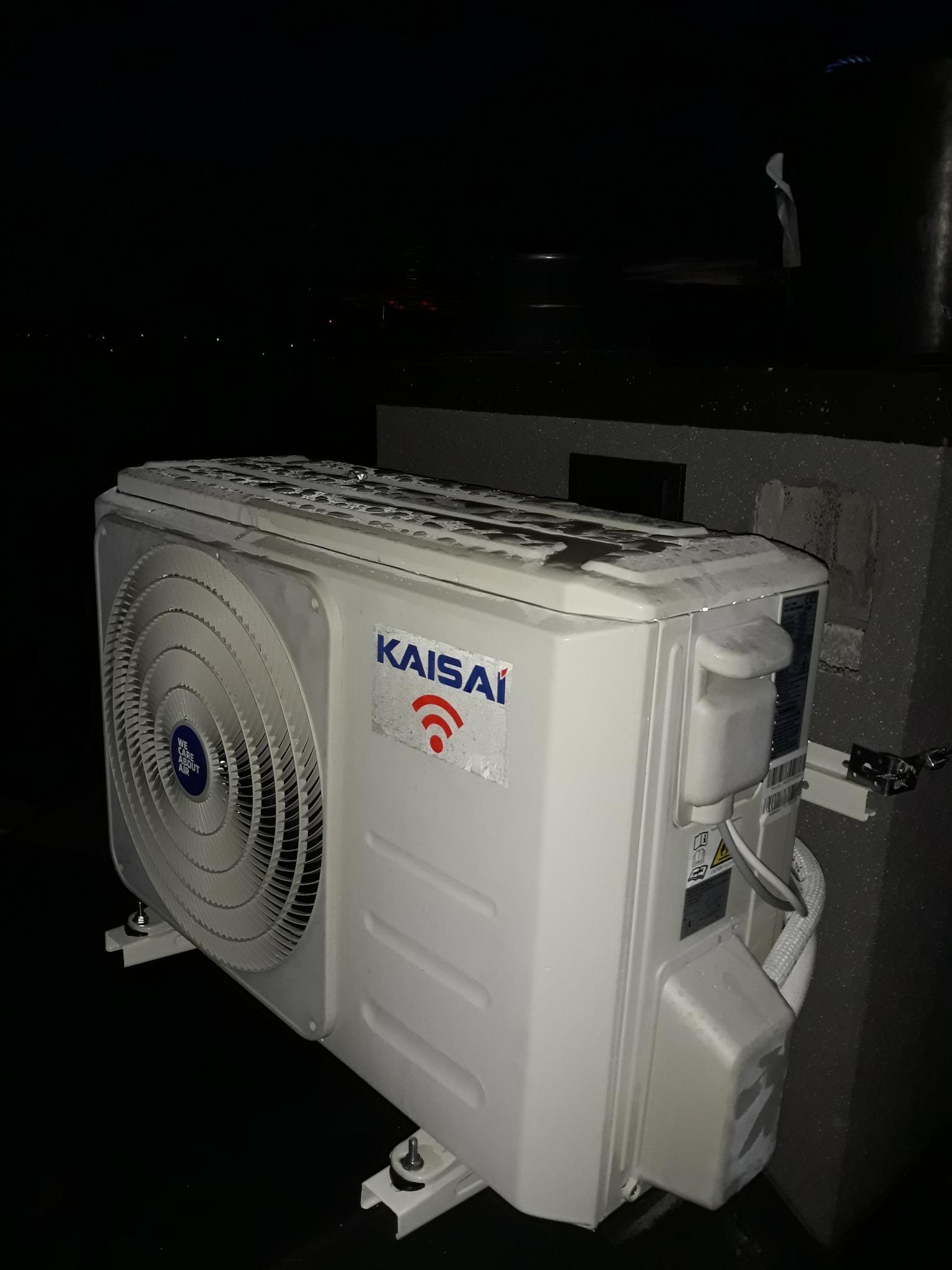 Zewnętrzna jednostka klimatyzacji Kaisai zamontowana na wspornikach, widoczne ślady wilgoci na obudowie, nocne ujęcie.