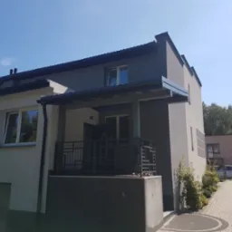 Dwukondygnacyjny dom jednorodzinny z szarą elewacją i ciemnym dachem, widok z dołu na balkon z metalową balustradą o geometrycznym wzorze oraz fragment dachu z rynnami.