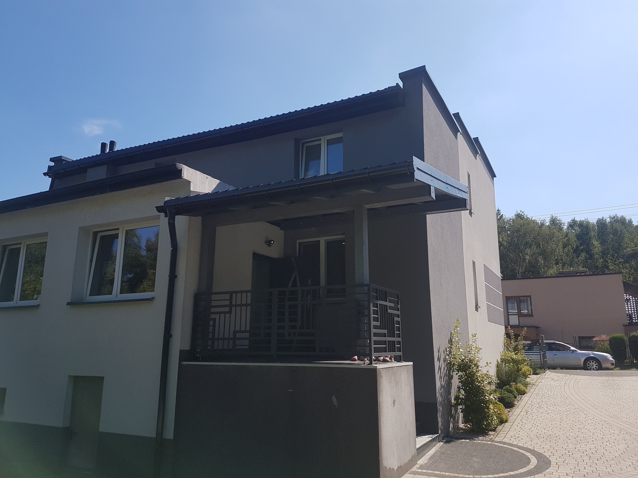Dwukondygnacyjny dom jednorodzinny z szarą elewacją i ciemnym dachem, widok z dołu na balkon z metalową balustradą o geometrycznym wzorze oraz fragment dachu z rynnami.