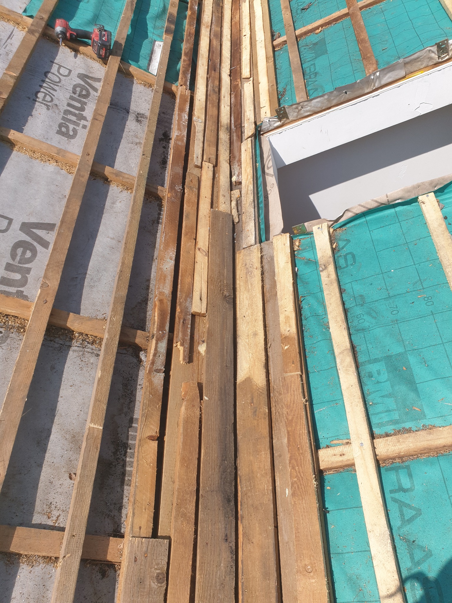 Widok z góry na częściowo deskowany dach z użyciem zielonej membrany i wkrętarki akumulatorowej. Etap prac przed ostatecznym pokryciem.