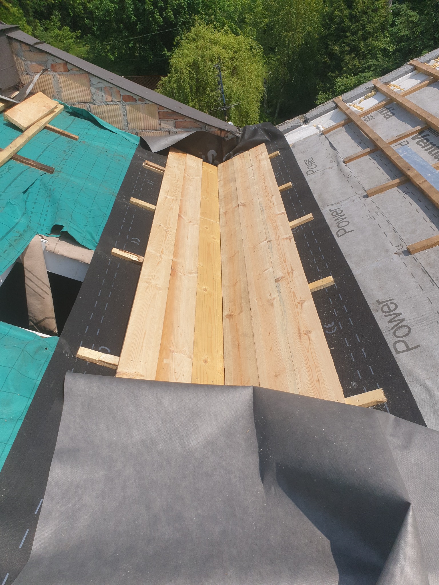 Dach w trakcie budowy: widoczne deski, membrana dachowa i łaty. Elementy konstrukcji dachu w zbliżeniu, zielona membrana i szara folia wiatroizolacyjna.