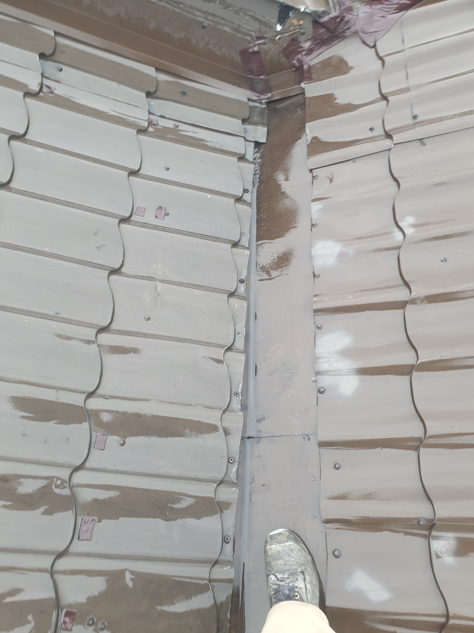 Widok z góry na fragment dachu z blachy trapezowej przed naprawą. Widoczne ślady korozji i zabrudzenia. But pracownika w prawym dolnym rogu kadru.