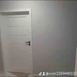 Fil Bud - Nowoczesne, białe drzwi wewnętrzne z poziomymi frezami i minimalistyczną klamką na tle szarej ściany, widoczne fragmenty podłogi zabezpieczonej kartonem.