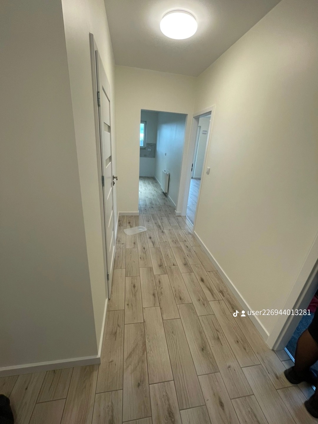 Wnętrze korytarza z panelami podłogowymi imitującymi drewno, świeżo pomalowanymi ścianami w odcieniach bieli i widocznymi drzwiami po lewej stronie. W głębi widoczne pomieszczenie z grzejnikiem.