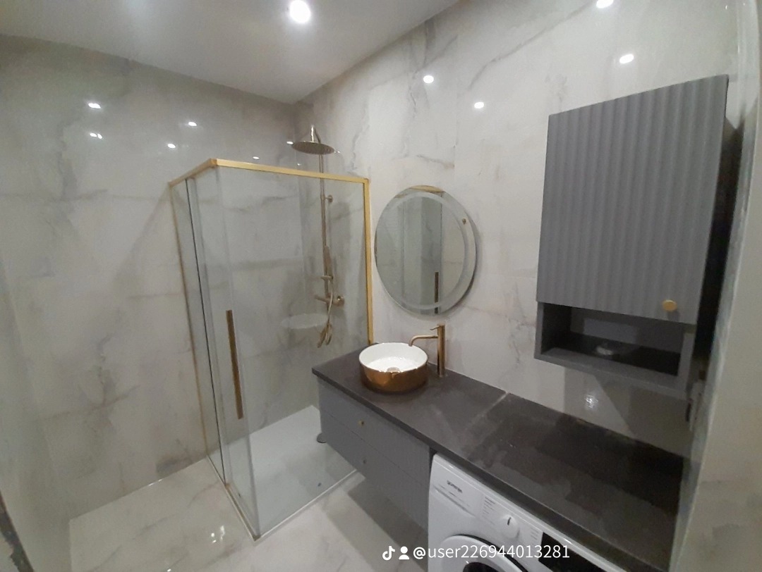 Nowoczesna łazienka z kabiną prysznicową ze złotymi ramami, okrągłym lustrem z podświetleniem LED, umywalką w kolorze złota, szarą szafką z umywalką, szarą szafką wiszącą z ryflowanym frontem...