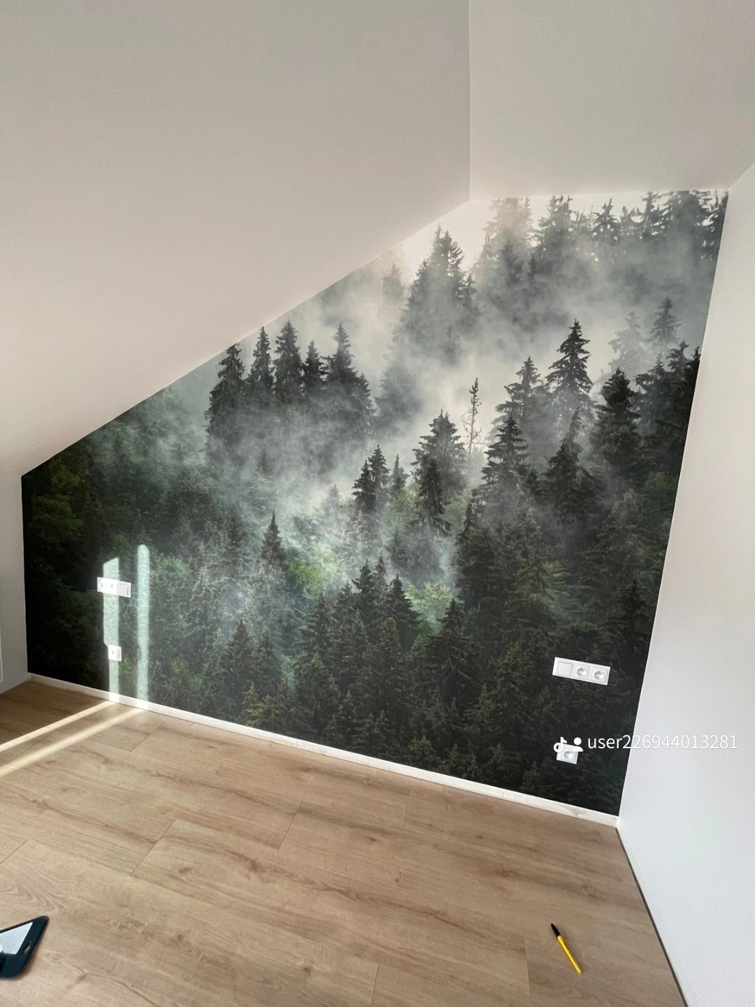 Ściana w pokoju na poddaszu pokryta fototapetą z motywem mglistego lasu iglastego, drewniana podłoga, widoczne gniazdka elektryczne.