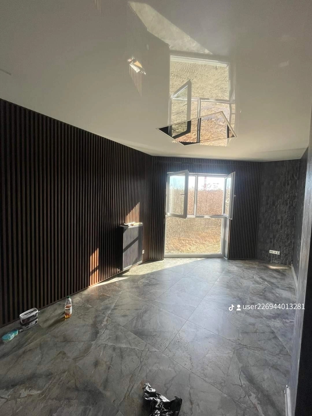 Wnętrze pokoju po remoncie z panelami ściennymi w kolorze ciemnego drewna, szarą marmurkową podłogą, grzejnikiem i nowoczesną lampą sufitową odbijającą światło, częściowo otwarte okno wpuszcza...