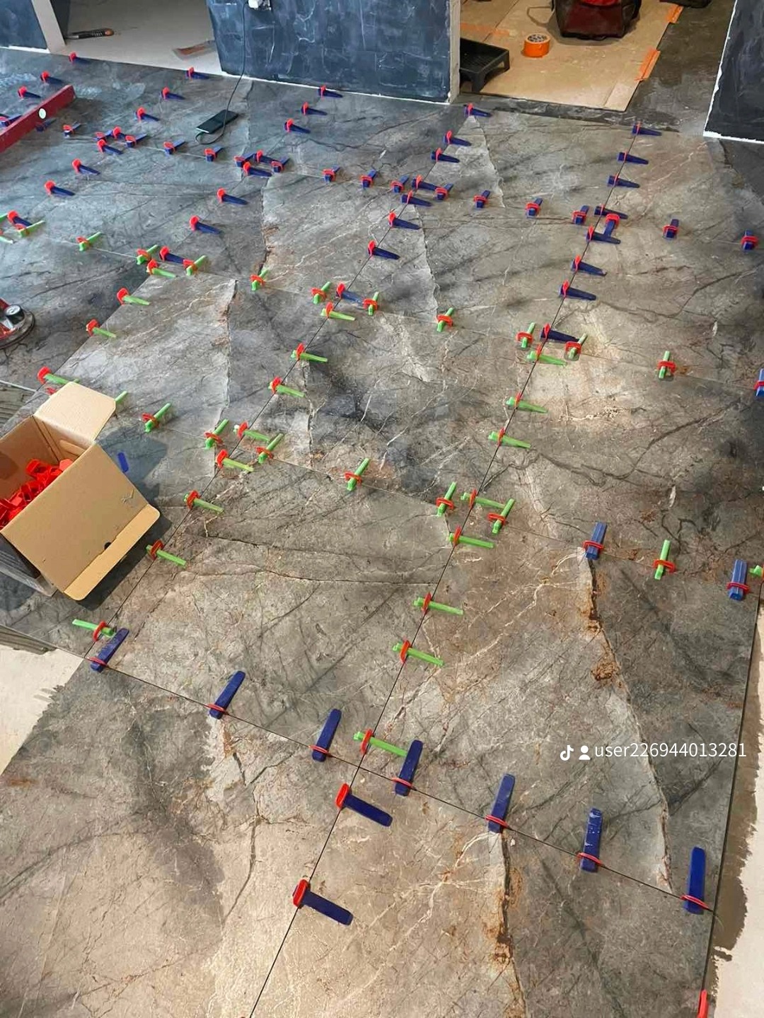 Układanie płytek podłogowych z użyciem systemu poziomowania: niebieskie i zielone klipsy wzdłuż czarnych sznurków na marmurowych płytach, karton z czerwonymi klinami w rogu.