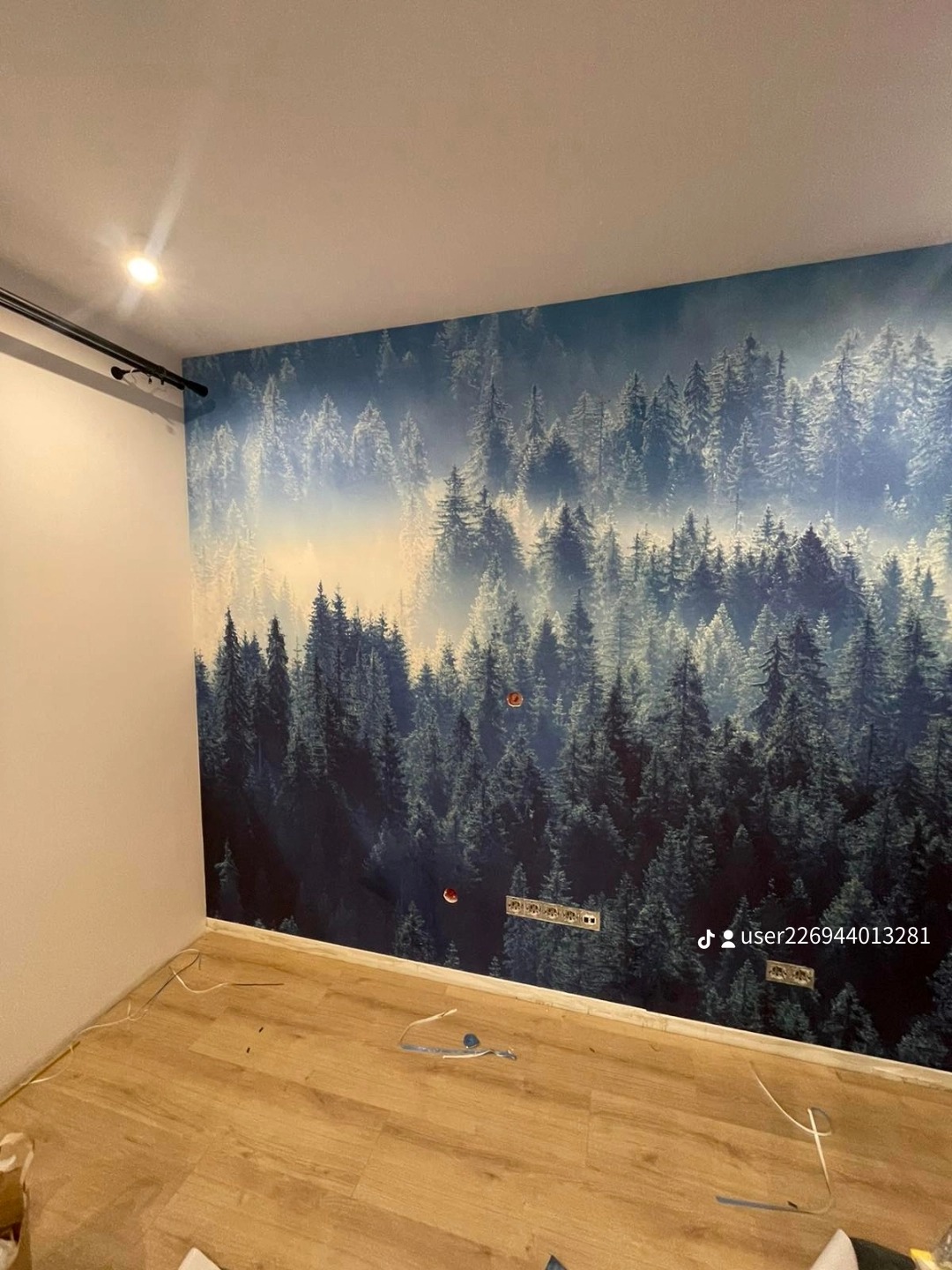 Ściana w pokoju z fototapetą przedstawiającą mglisty las iglasty, widoczne gniazdka elektryczne i wystające kable, podłoga wyłożona panelami.