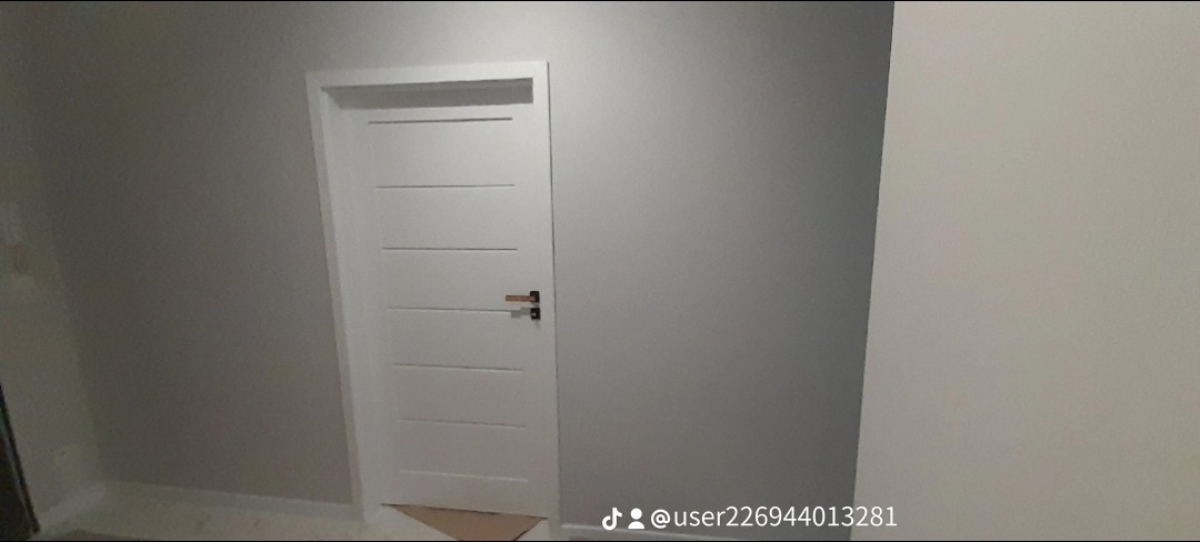 Nowoczesne, białe drzwi wewnętrzne z poziomymi frezami i minimalistyczną klamką na tle szarej ściany, widoczne fragmenty podłogi zabezpieczonej kartonem.